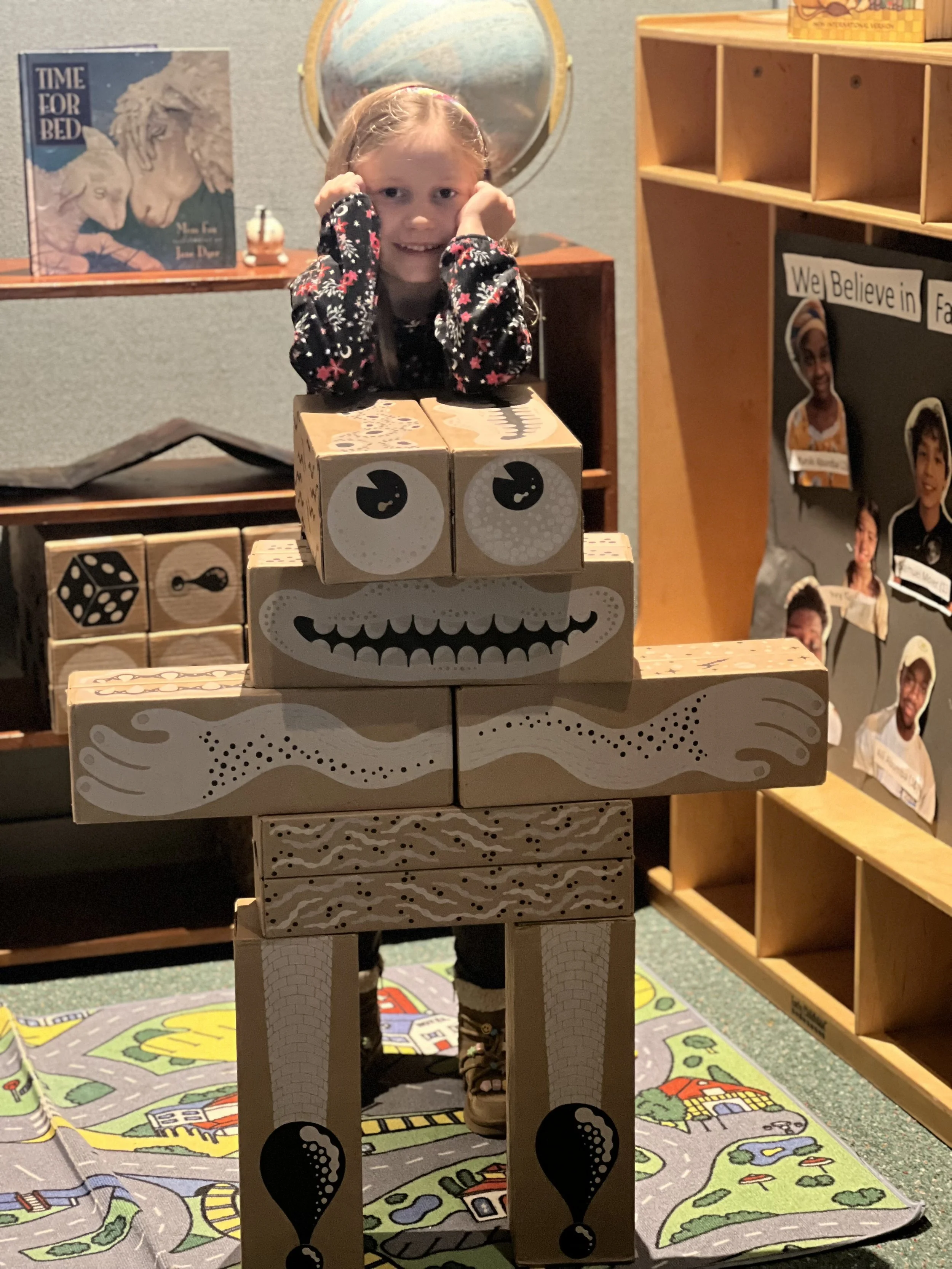 A young girl is smiling and resting her head on her hands behind a cardboard robot made from painted boxes with a happy face, long legs, and arms, in a playroom with educational posters and toy shelves.