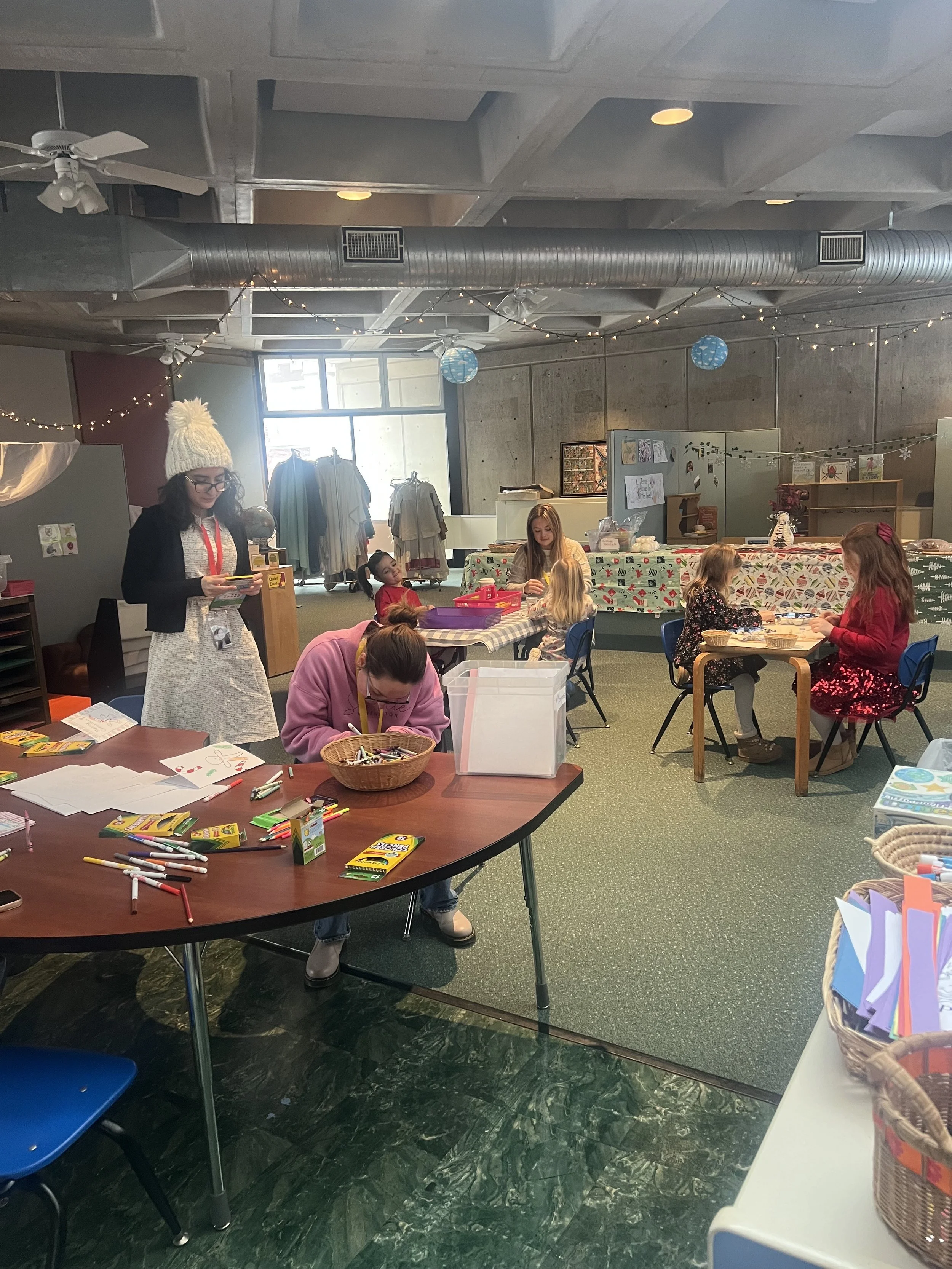 Children engaging in arts and crafts activities at tables in a decorated indoor space, with adults supervising and some holiday-themed decorations visible.