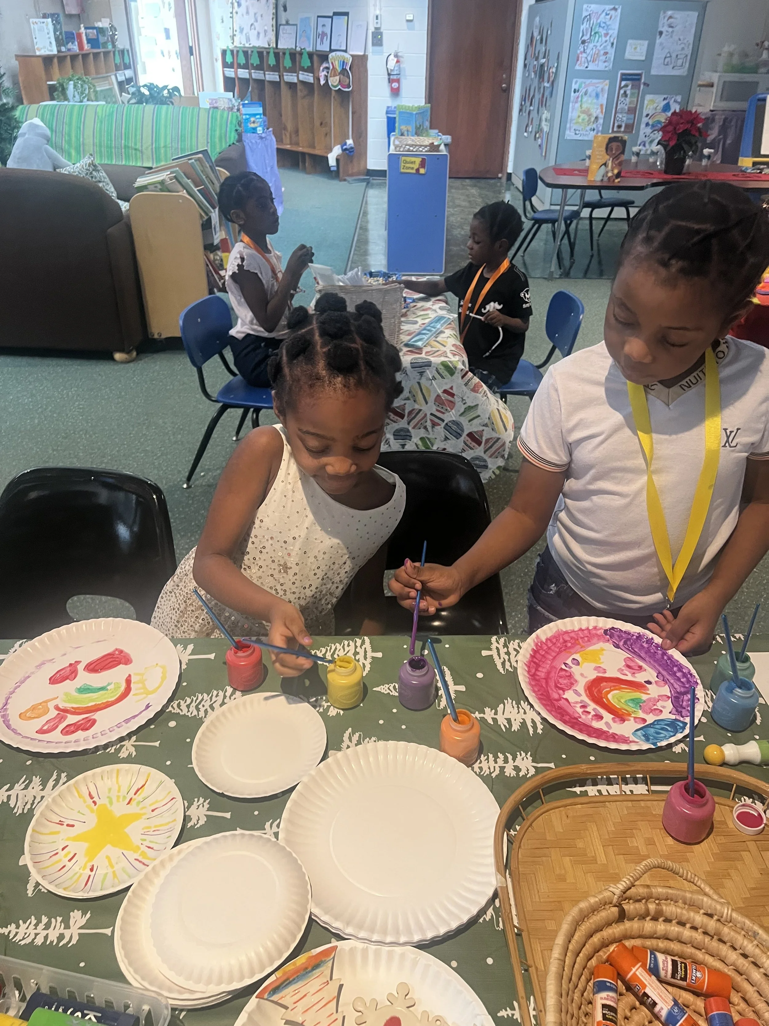 Children painting with colorful paints at a table in a classroom decorated for Christmas.