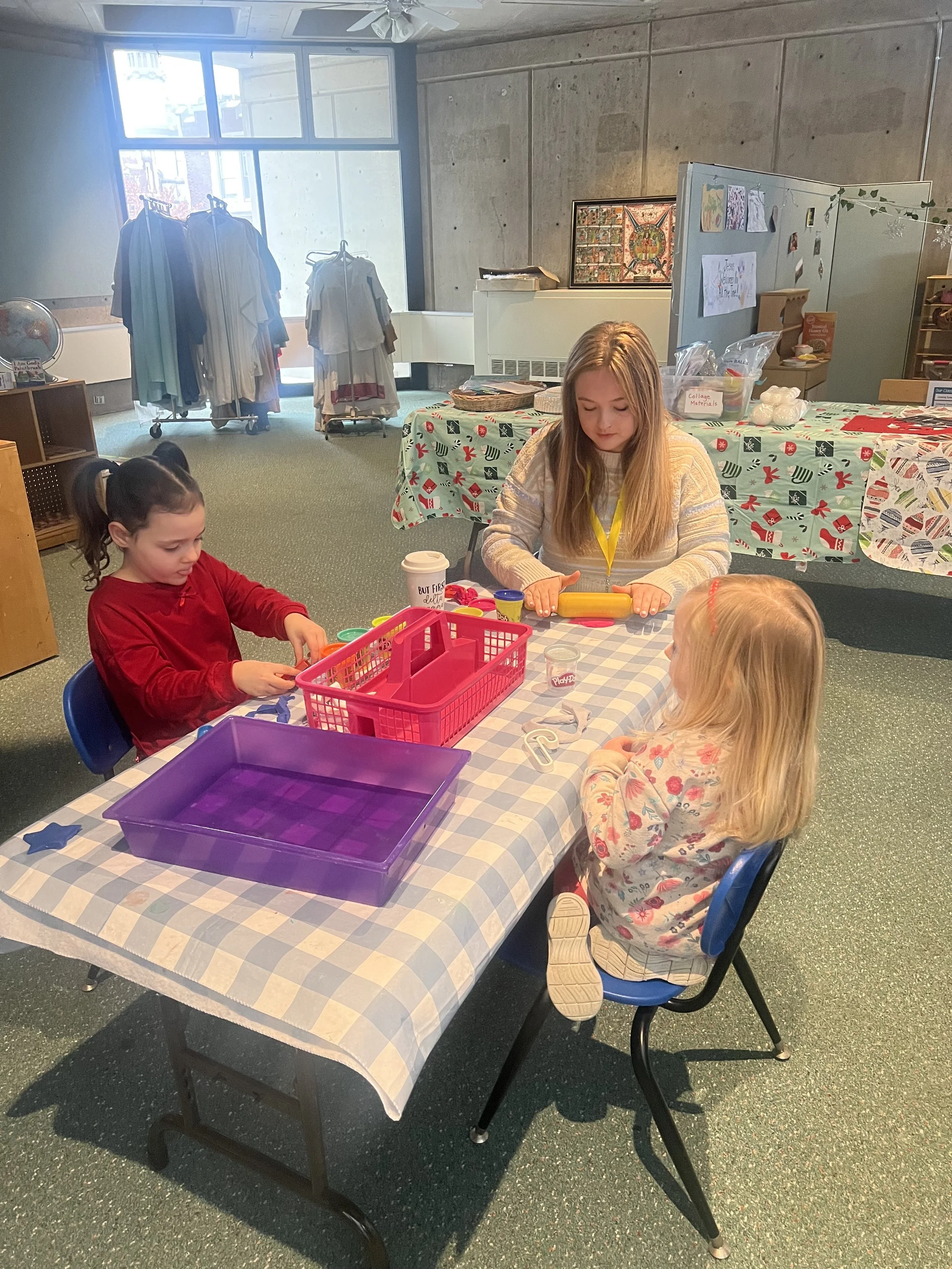 Three young girls are sitting at a table in a room with a large window, engaging in a craft activity with playdough and tools.