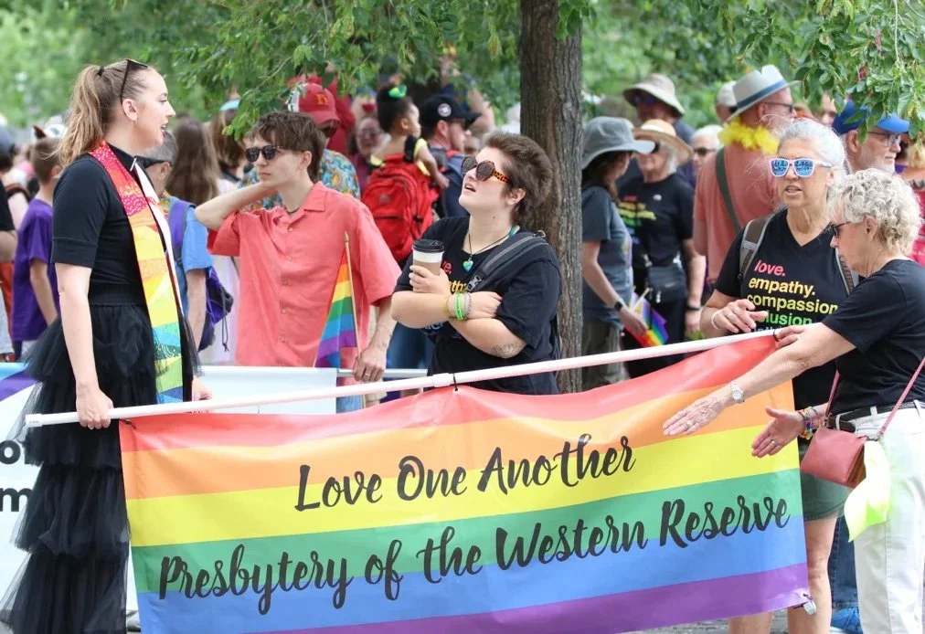 Group of people participating in a Pride parade holding a rainbow flag that reads, "Love One Another, Presbytery of the Western Reserve." Some individuals wearing rainbow-colored accessories and sunglasses, celebrating LGBTQ+ pride.
