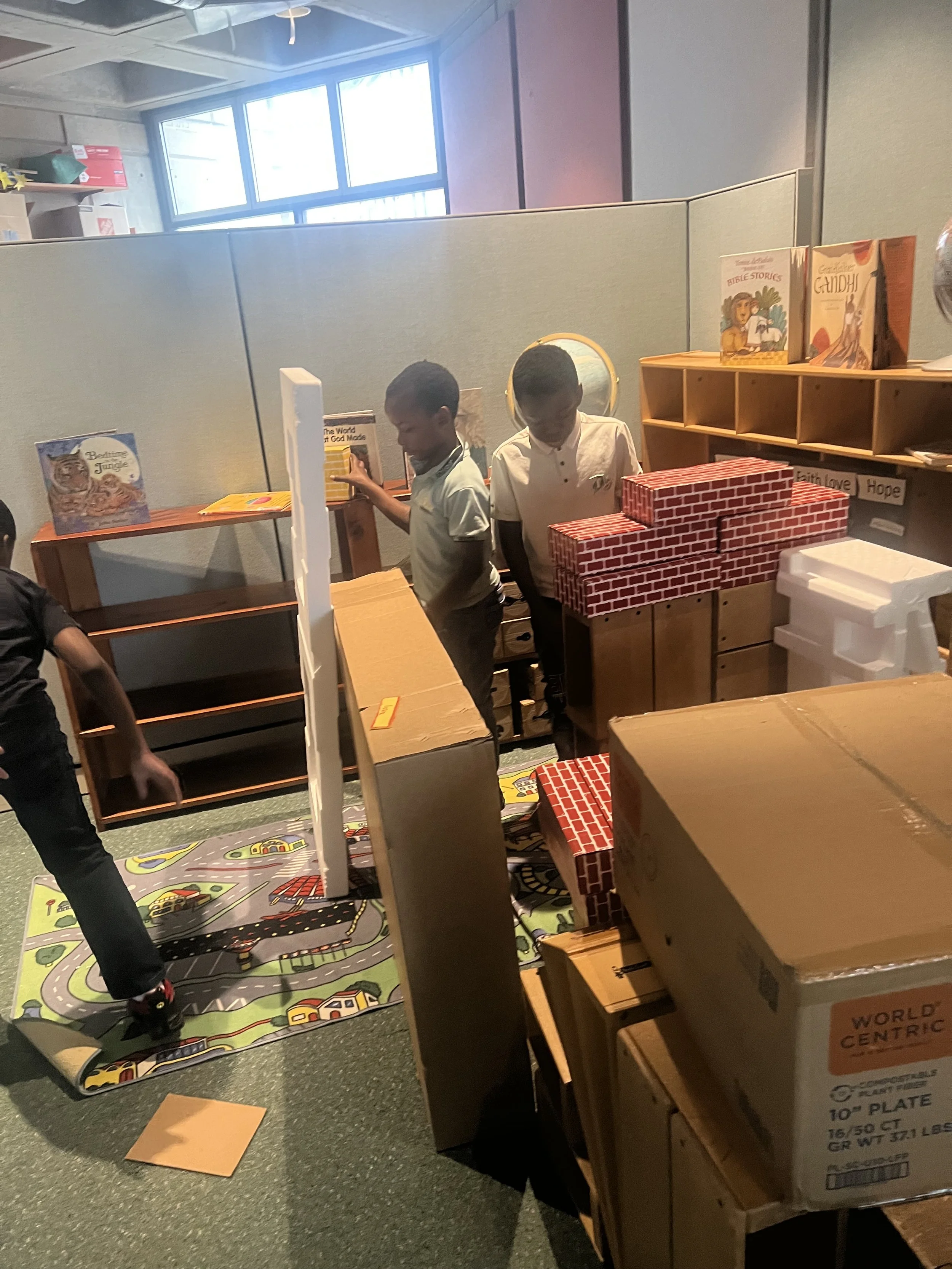Children standing in a room with bookshelves and cardboard boxes, some decorated like brick walls, on a colorful play mat with roads and houses.