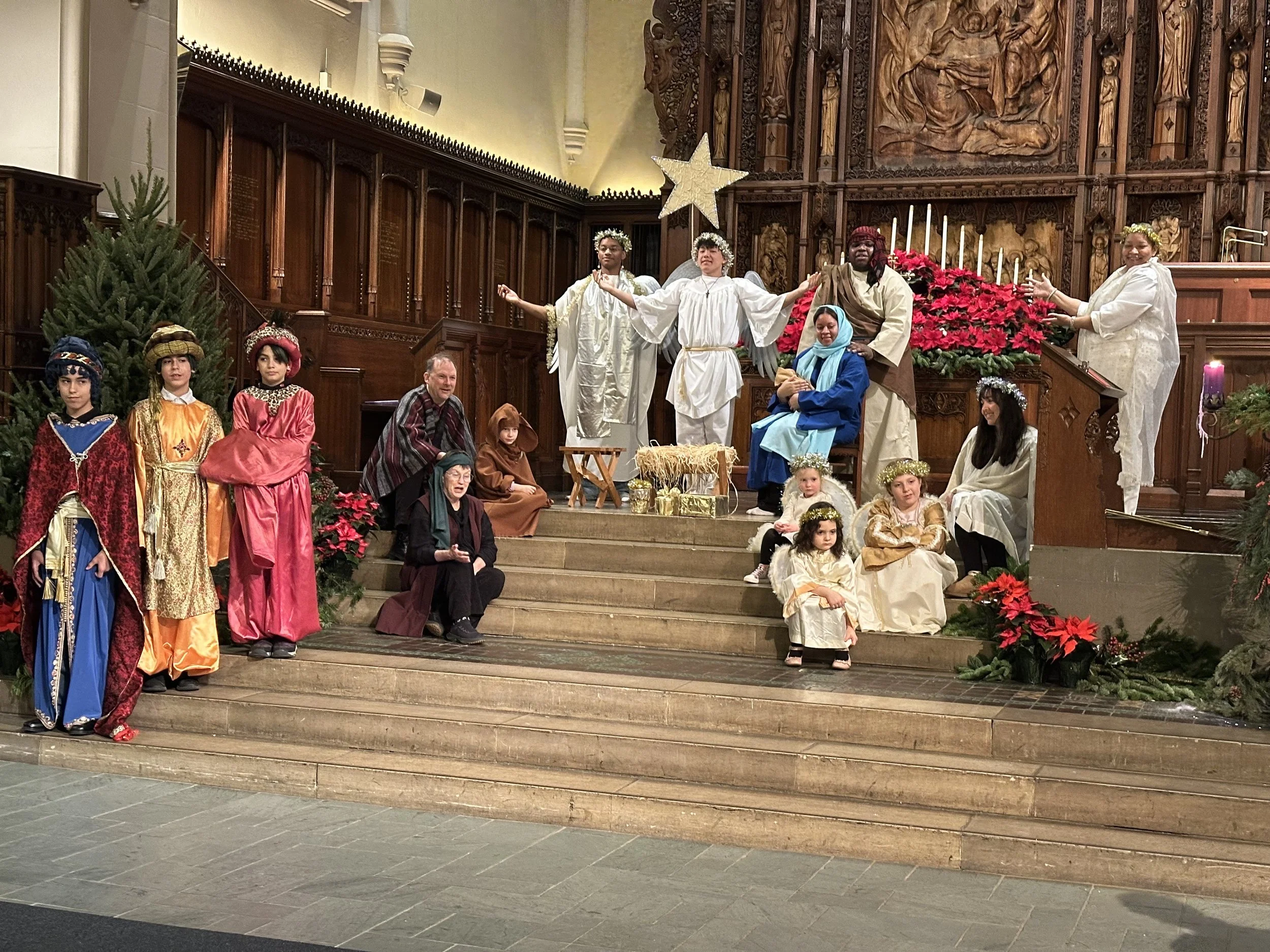 A Christmas play depicting the Nativity scene with children and adults dressed as Mary, Joseph, angels, Wise Men, shepherds, and animals inside a church with wooden carvings and poinsettias.