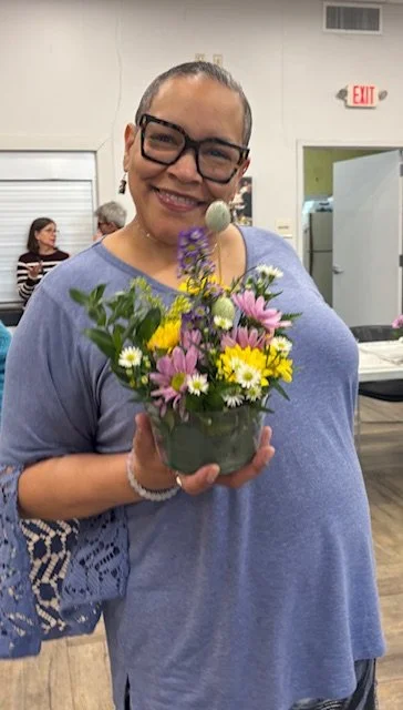 A smiling woman with glasses holding a bouquet of colorful flowers in a room with a few other people in the background.