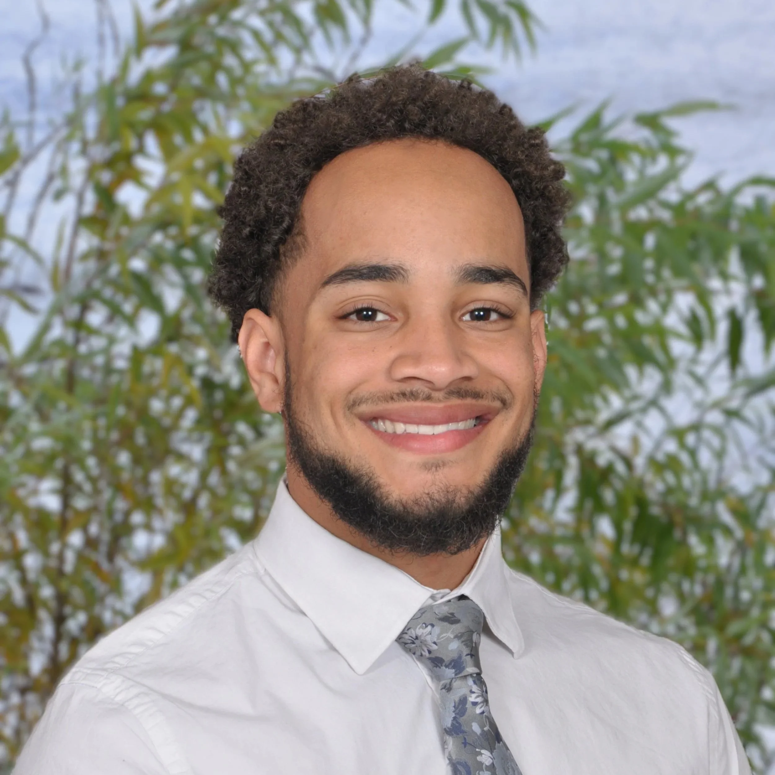 A smiling young man with curly hair and a beard, wearing a white shirt and a patterned gray tie, standing outdoors in front of green foliage.