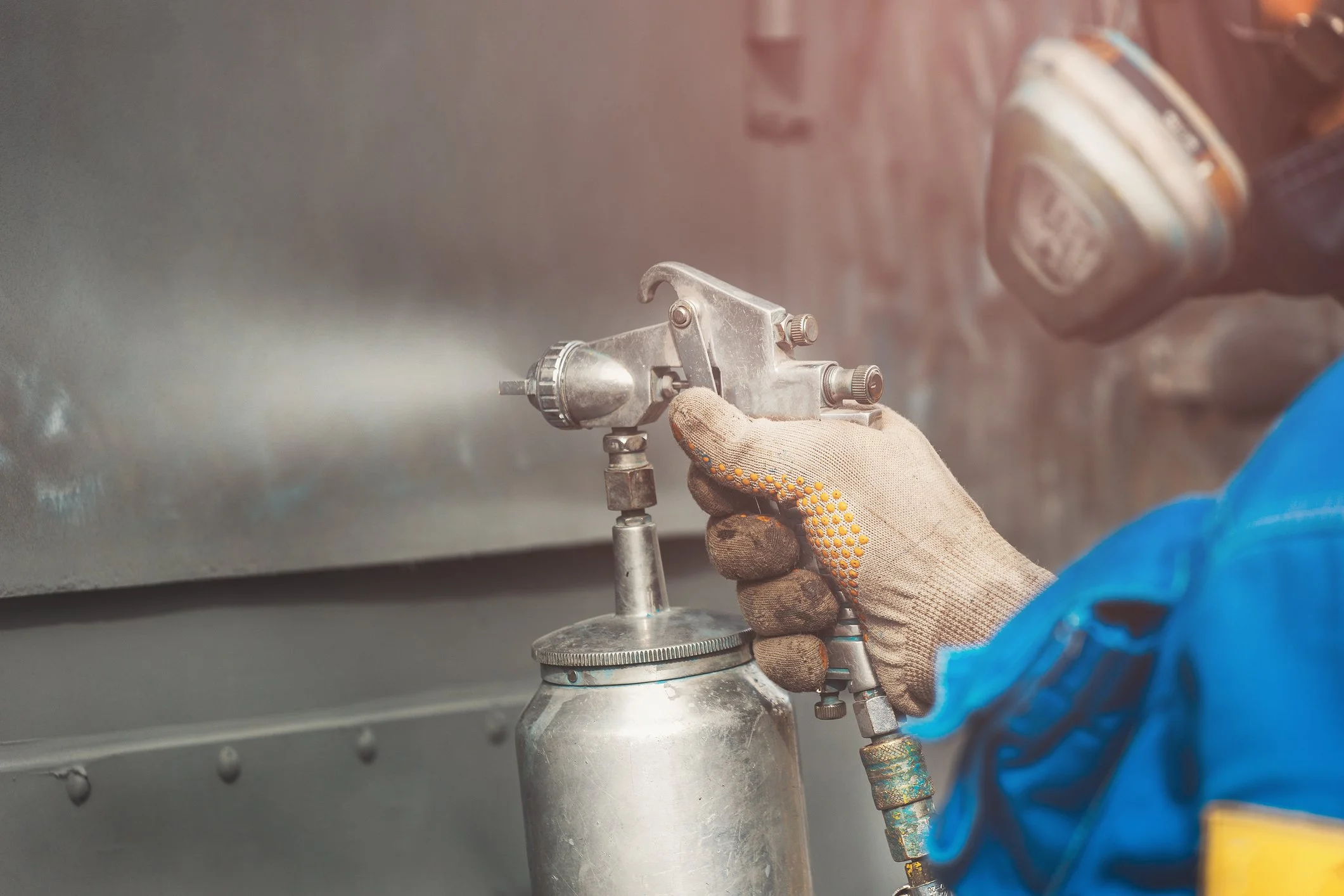 A worker wearing gloves and a blue uniform filling a tank or container with a liquid using a spray gun attached to a hose.