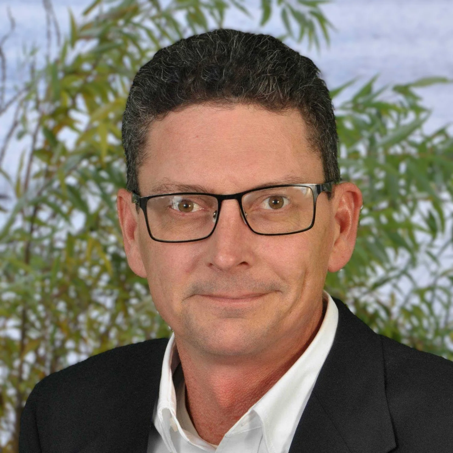 A man with short curly dark hair and glasses, dressed in a black suit and white shirt, standing in front of a leafy green background.