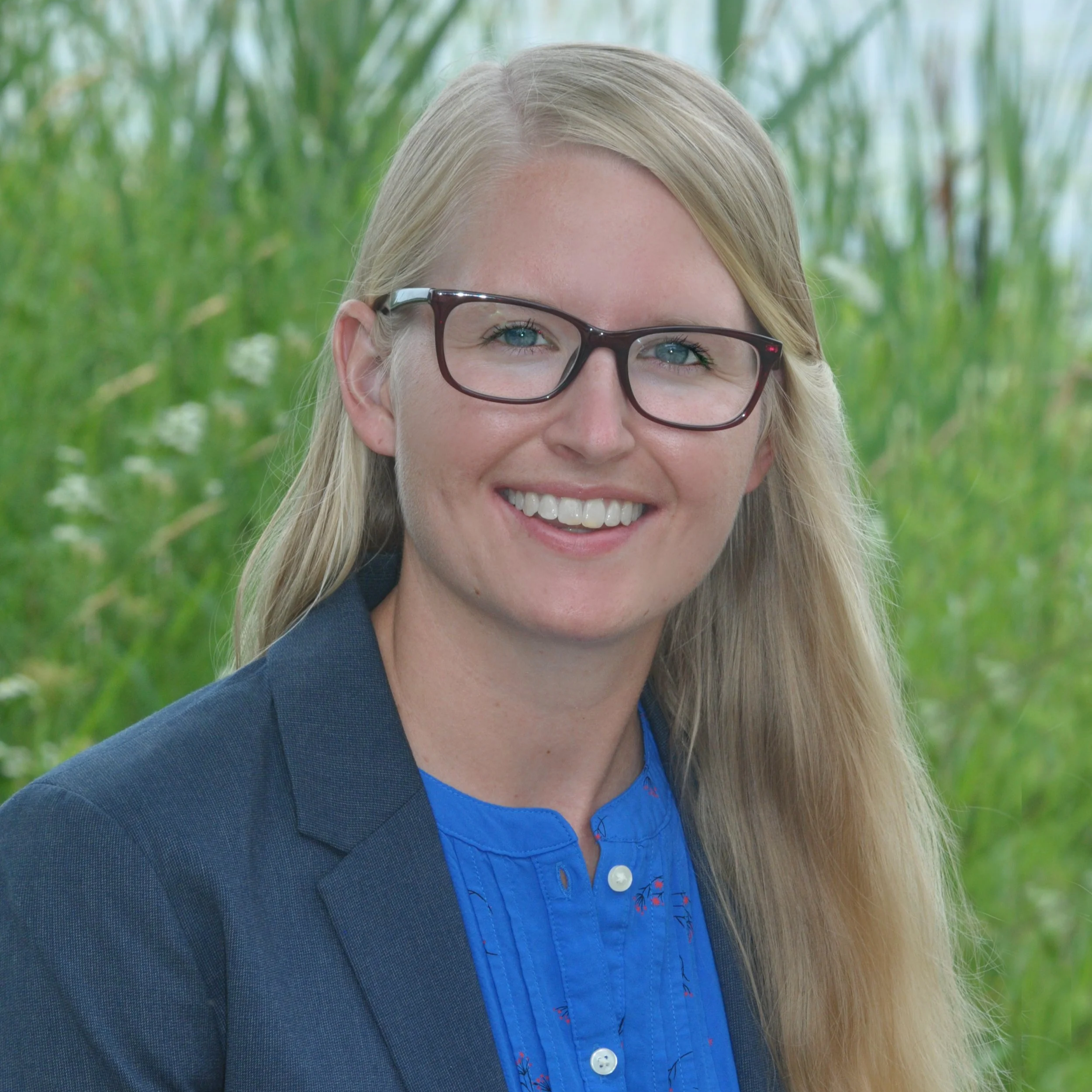 A smiling woman with blonde hair and glasses, wearing a blue blouse and a dark blazer, standing outdoors with tall green grass in the background.