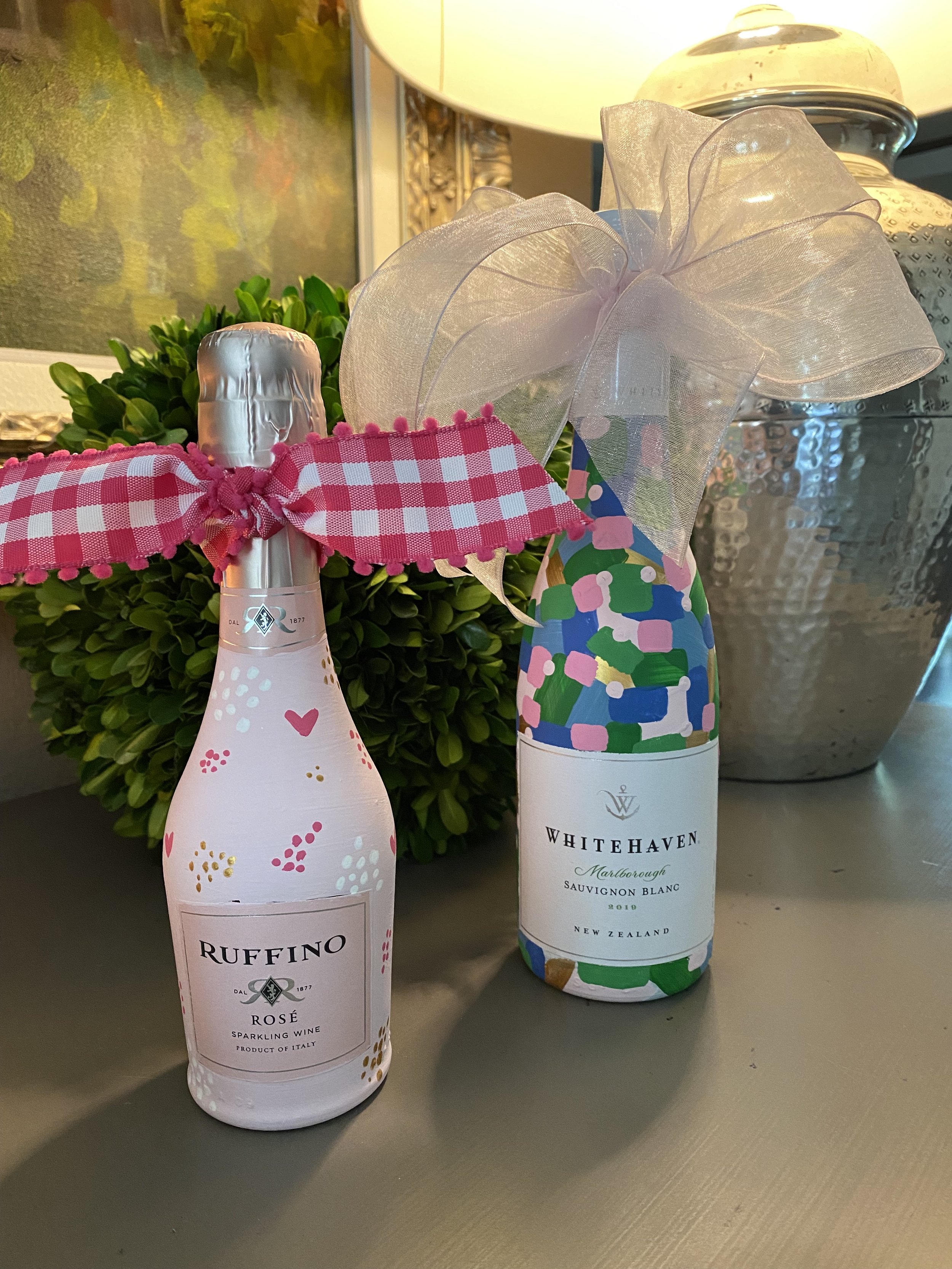 Two wine bottles, hand-painted in colorful designs. One is blush pink with tiny bubbles and a hot pink bow, the other is pink, green & blue with a blush bow.