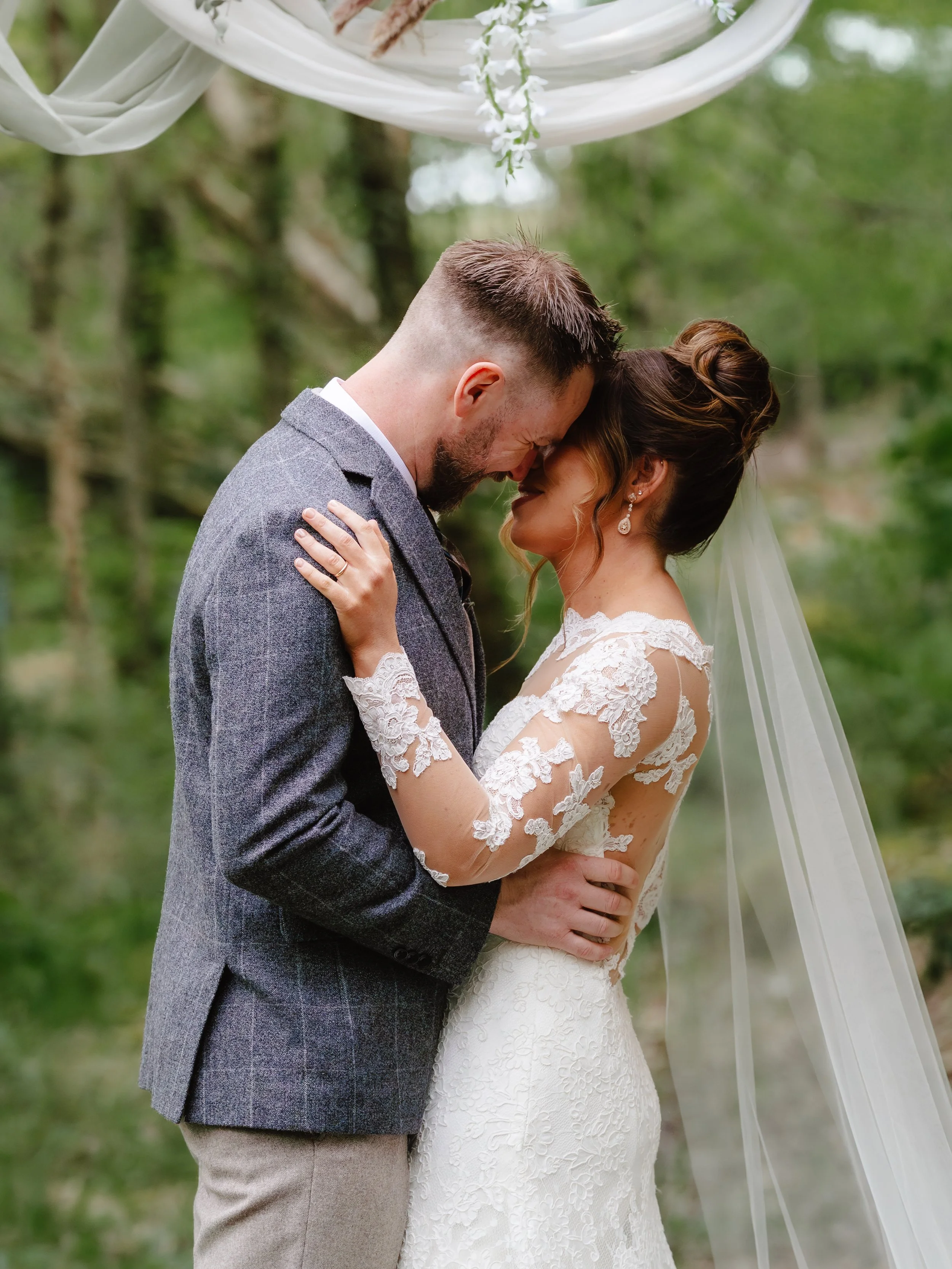 A bride and groom embrace during their wedding ceremony outdoors, touching foreheads with eyes closed, surrounded by greenery and decorated fabric and flowers overhead.