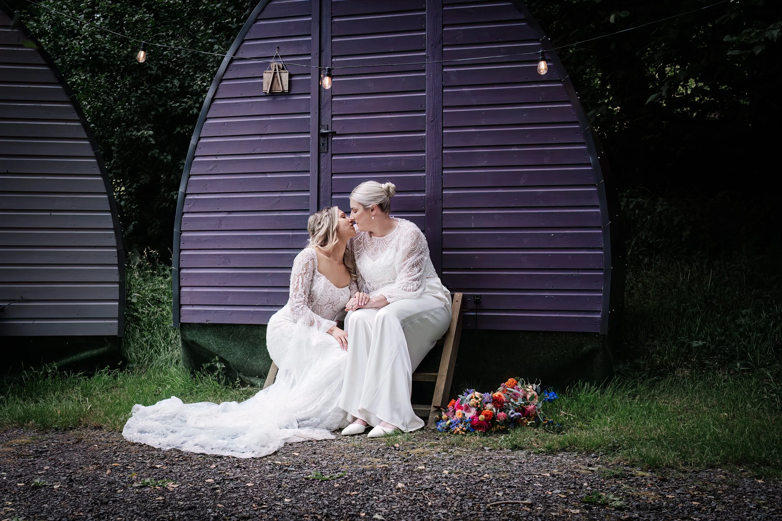 Two women, wearing white wedding dresses, sharing a kiss while seated outdoors on a small wooden bench in front of purple wooden structures with string lights hanging above, and a colorful bouquet lying on the ground nearby.