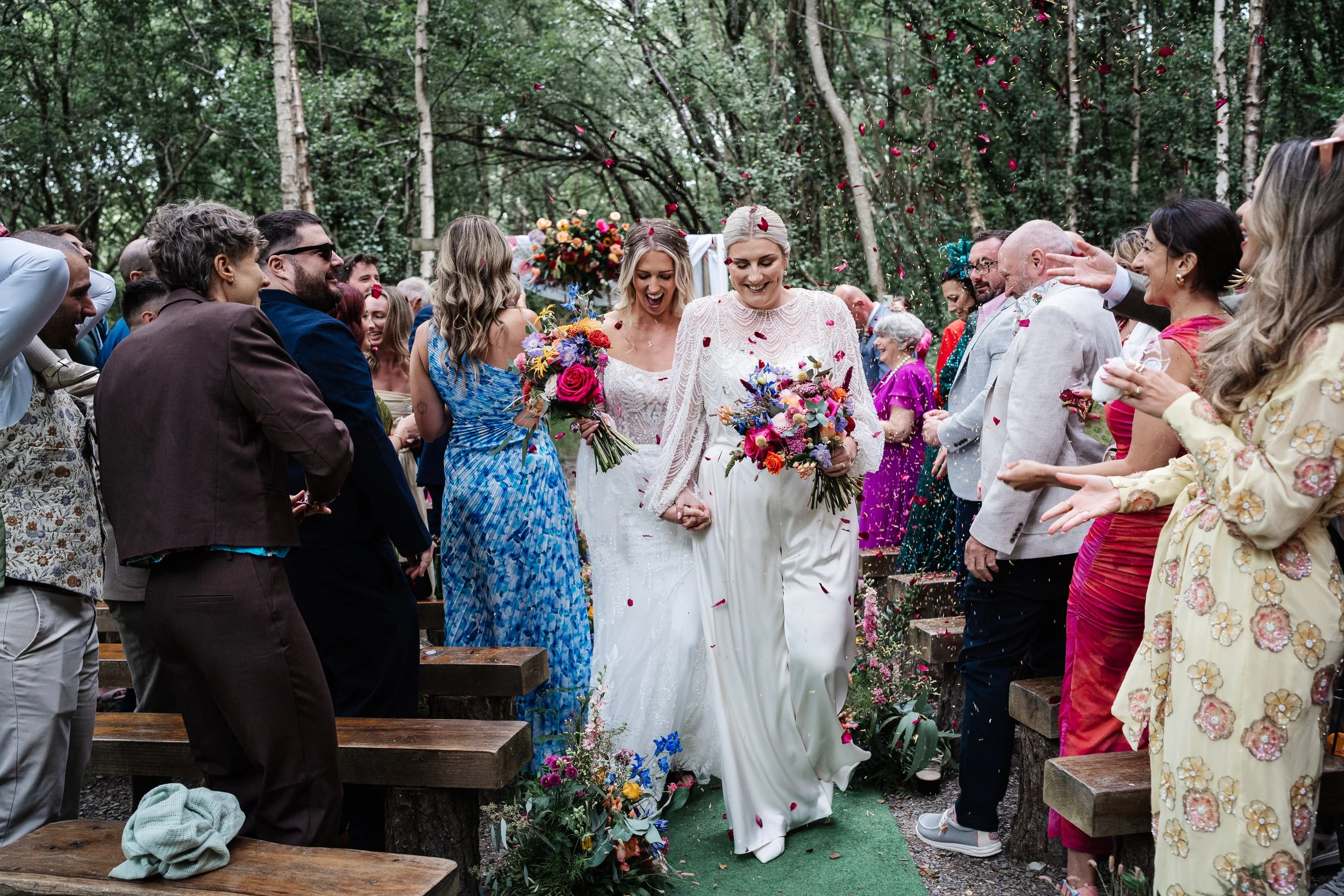 Two brides walking hand-in-hand down the aisle at an outdoor wedding ceremony, surrounded by guests throwing flower petals in the air, with trees in the background.