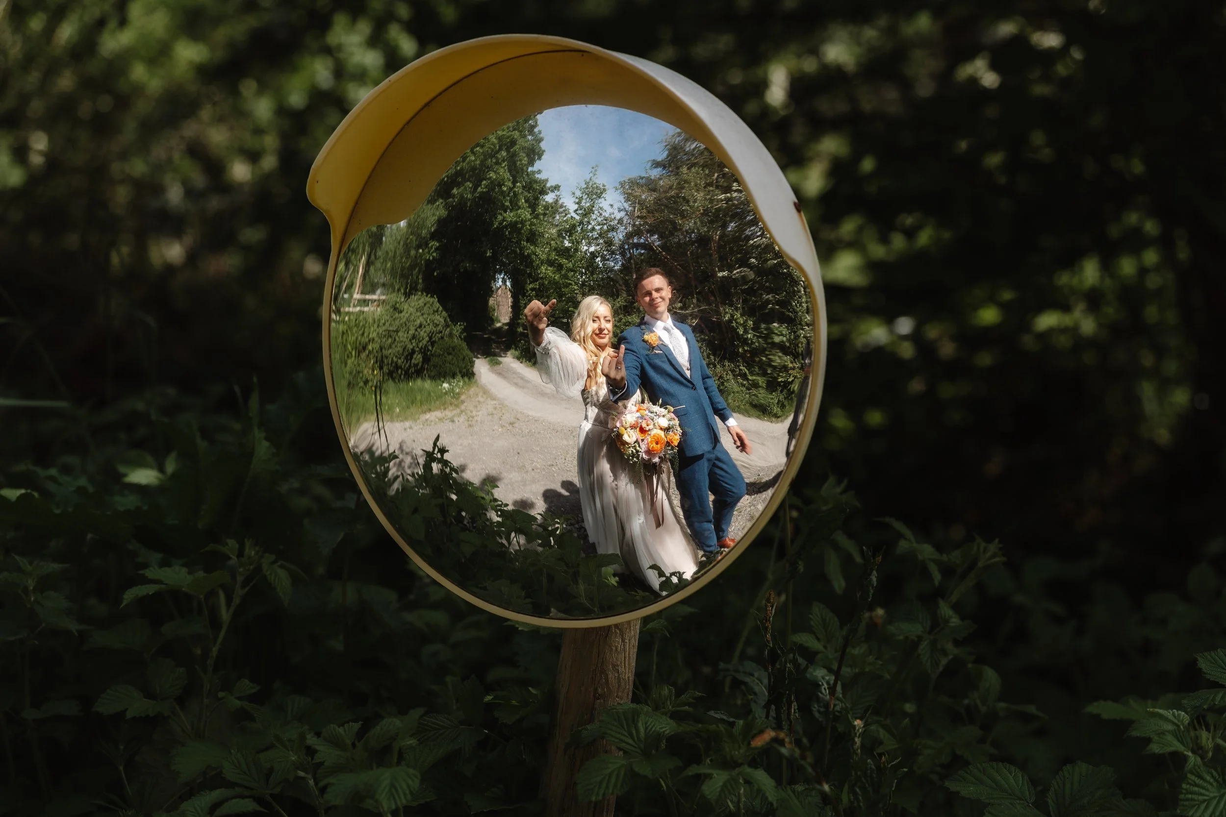 A reflection of a bride and groom in a convex mirror outdoors on a sunny day, surrounded by green trees and foliage.