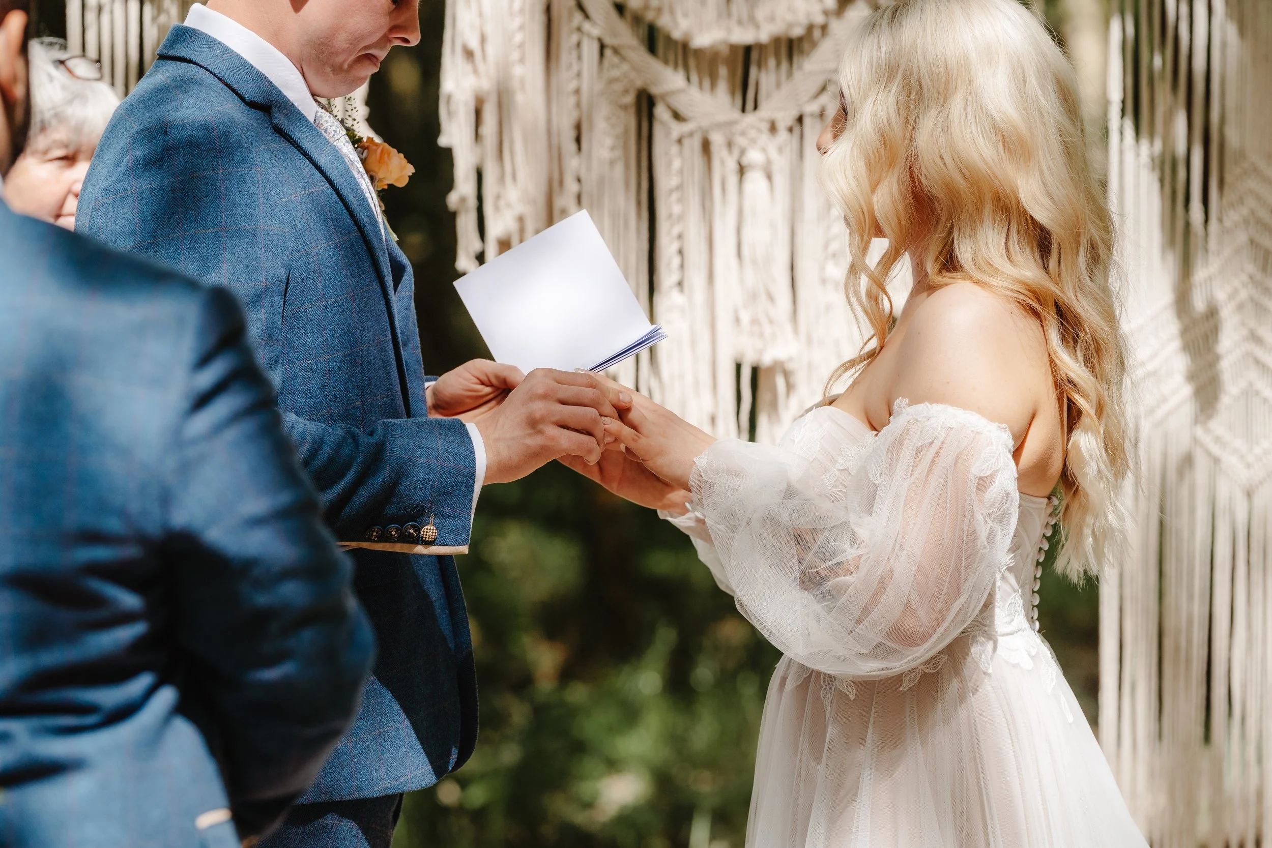 A couple exchanging wedding vows outdoors, with the groom reading from a paper and the bride holding his hands.