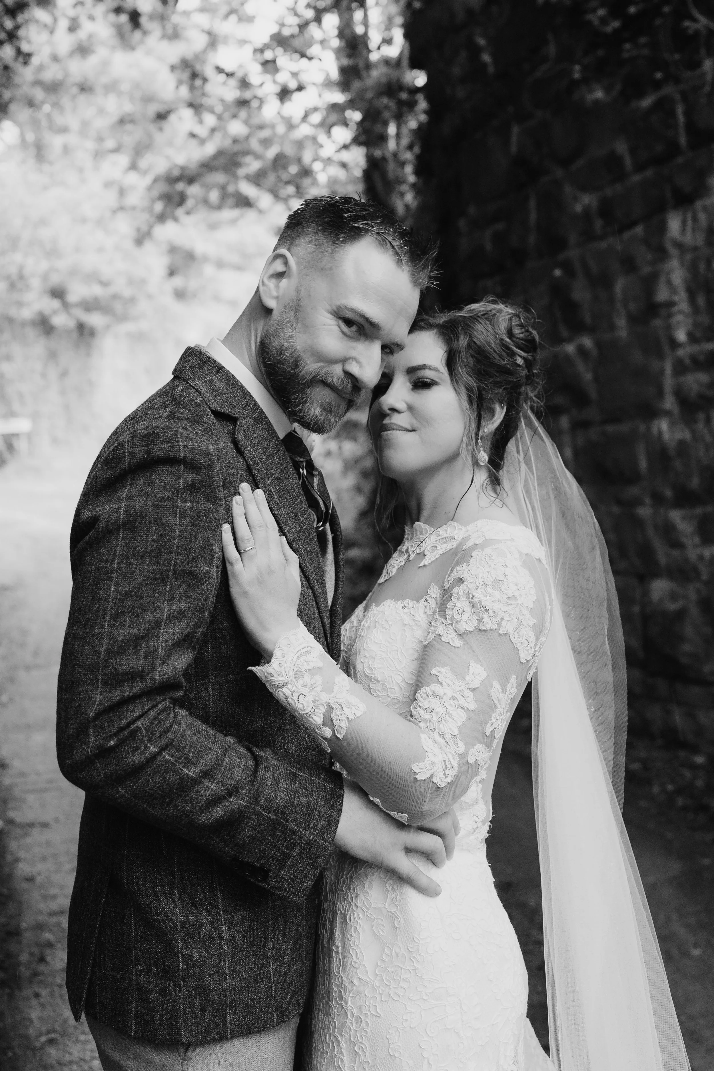 A black and white photo of a bride and groom embracing outdoors. The groom, with a beard and short hair, wears a textured suit jacket. The bride, with styled hair and earrings, wears a lace wedding dress and veil. They are smiling softly, close to ea