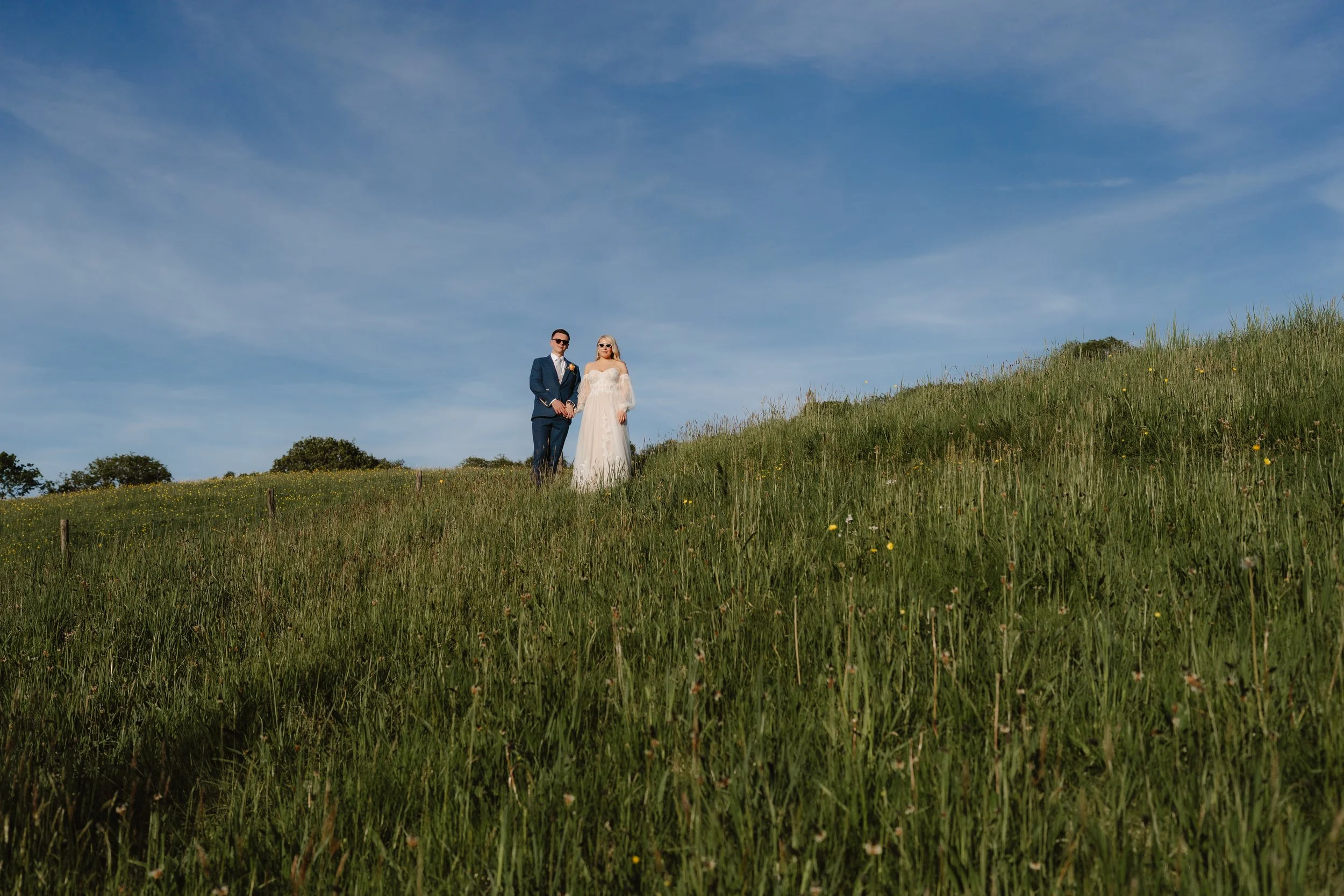 A bride and groom holding hands and standing in a grassy field on a sunny day with blue sky and some clouds.
