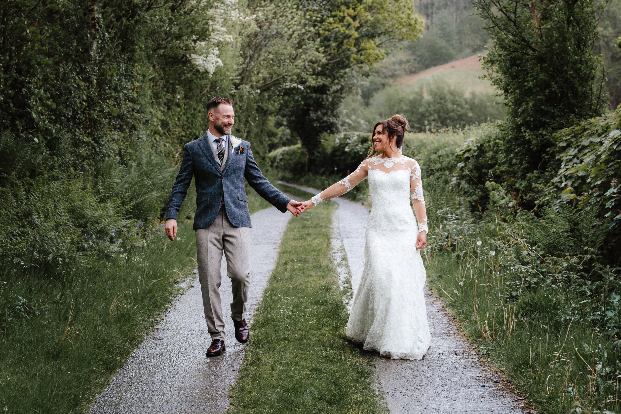 A bride and groom holding hands, walking on a gravel path in a lush green outdoor setting with trees and distant hills.