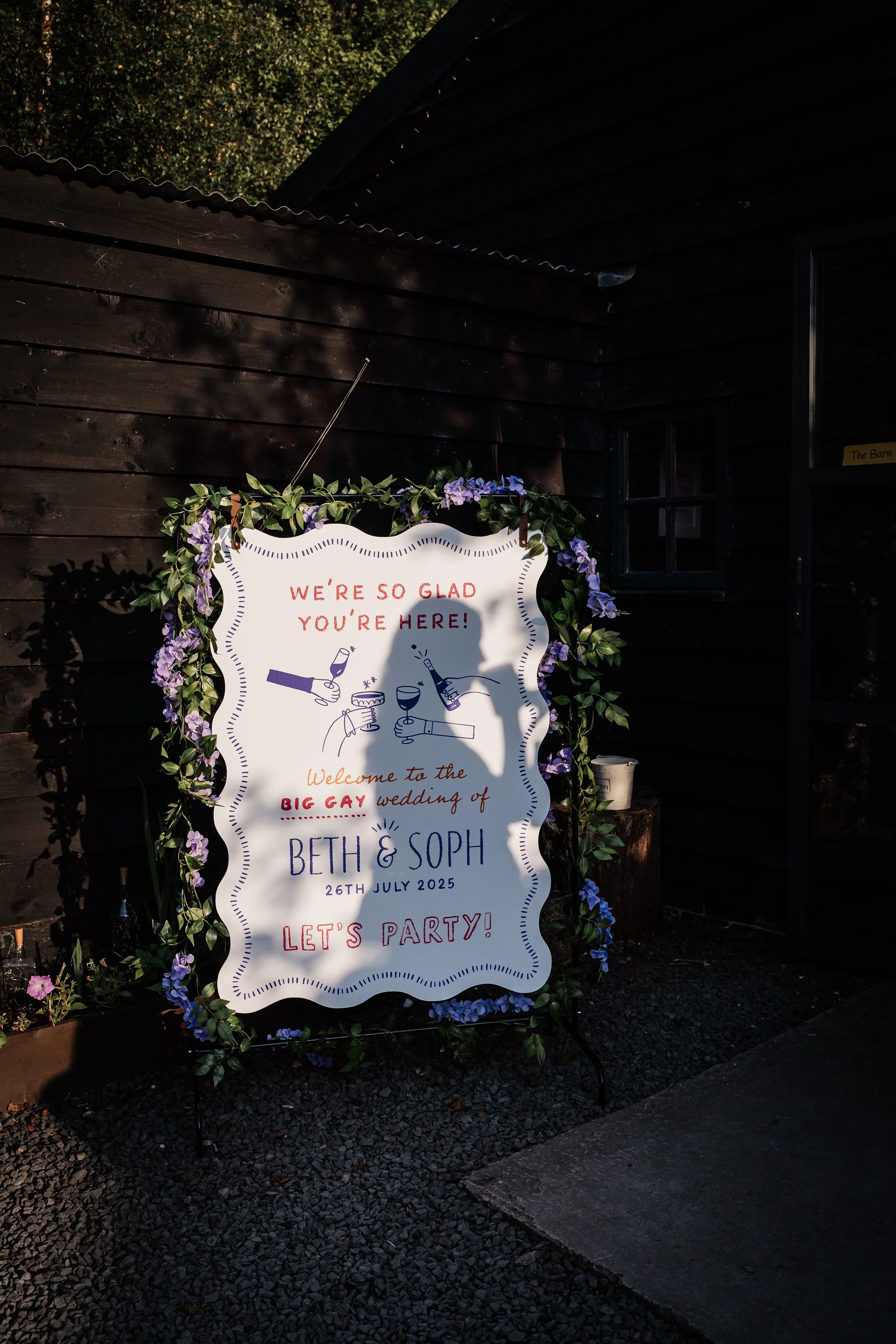 Welcoming sign decorated with purple flowers for Beth and Soph's wedding on July 26, 2025, celebrating with drinks and a party.