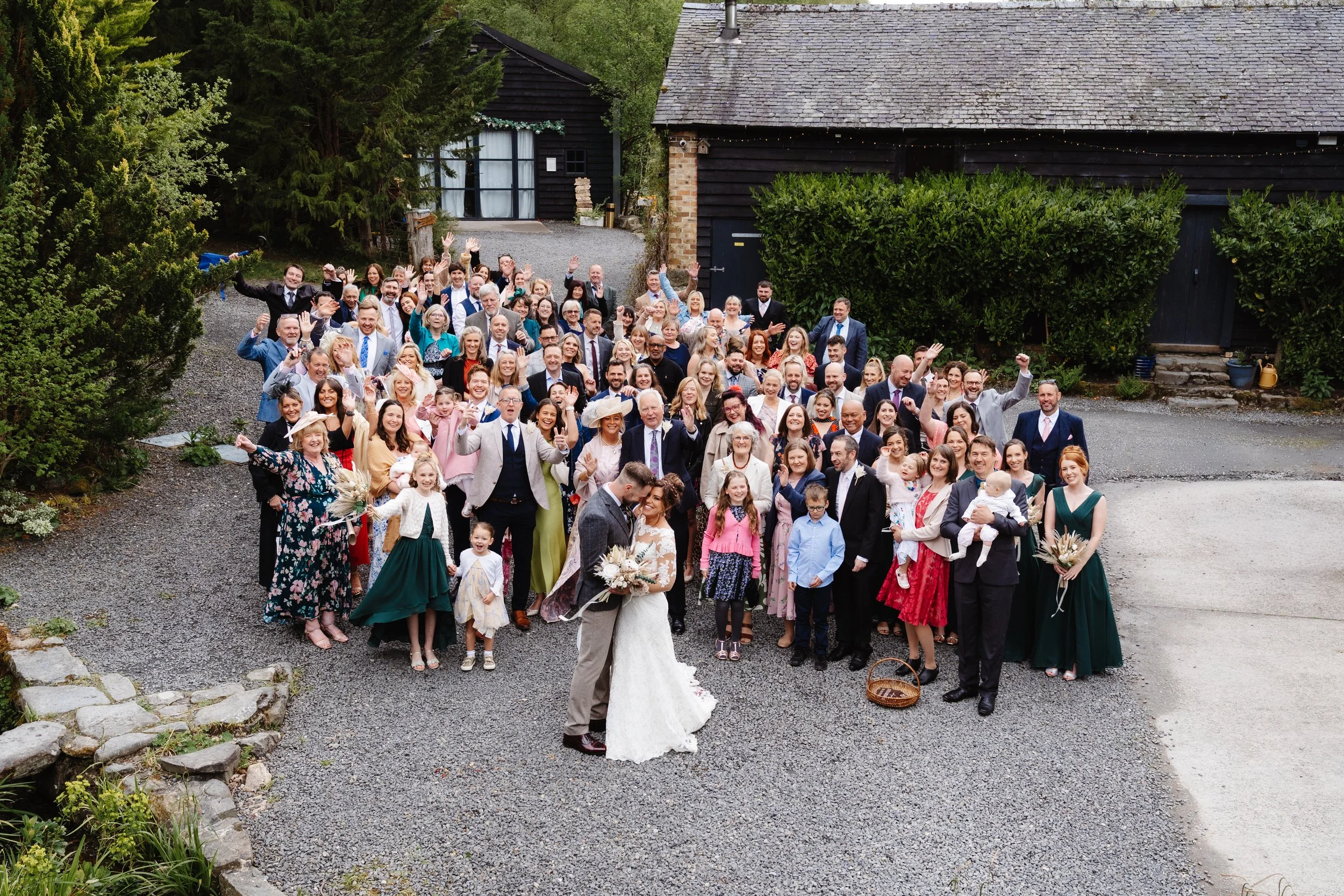 A large group of people gathered outdoors for a wedding, with the bride and groom in the front center. The group is smiling, waving, and celebrating in front of rustic buildings and lush greenery.