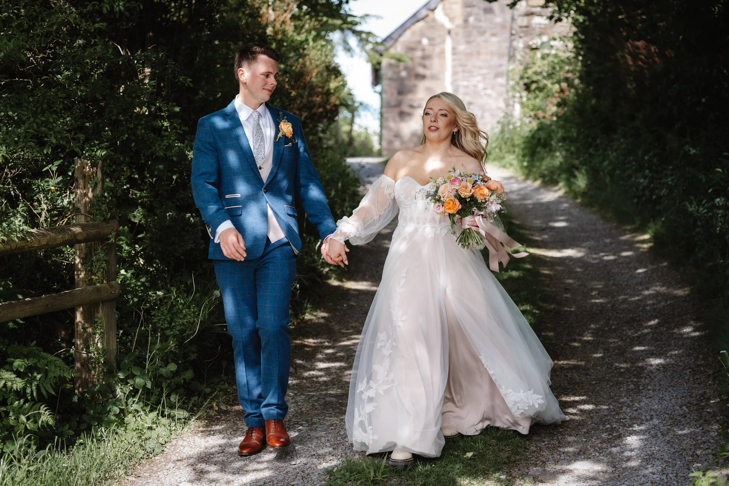 A bride and groom holding hands and walking outdoors on a sunny day, surrounded by greenery and trees, with a stone building in the background.