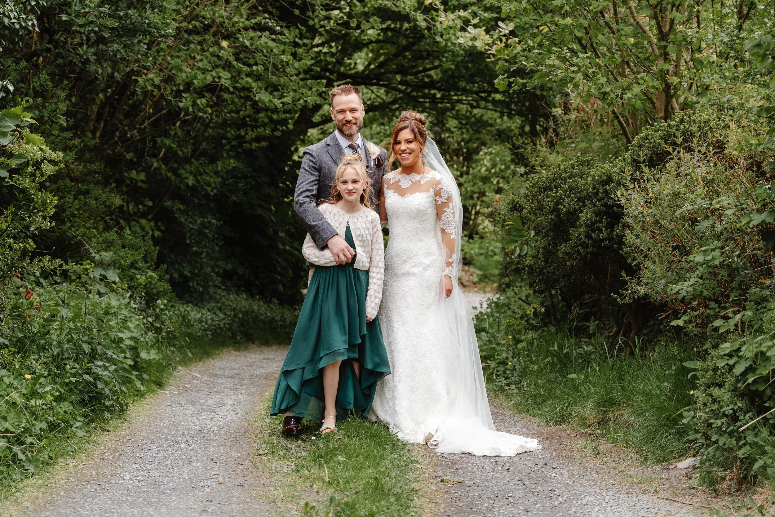 A bride with brown hair in an updo and wearing a white lace wedding dress with a veil stands beside a groom with a beard and short hair in a gray suit, along with a young girl with blonde hair in a green dress and white cardigan, on a wooded path sur