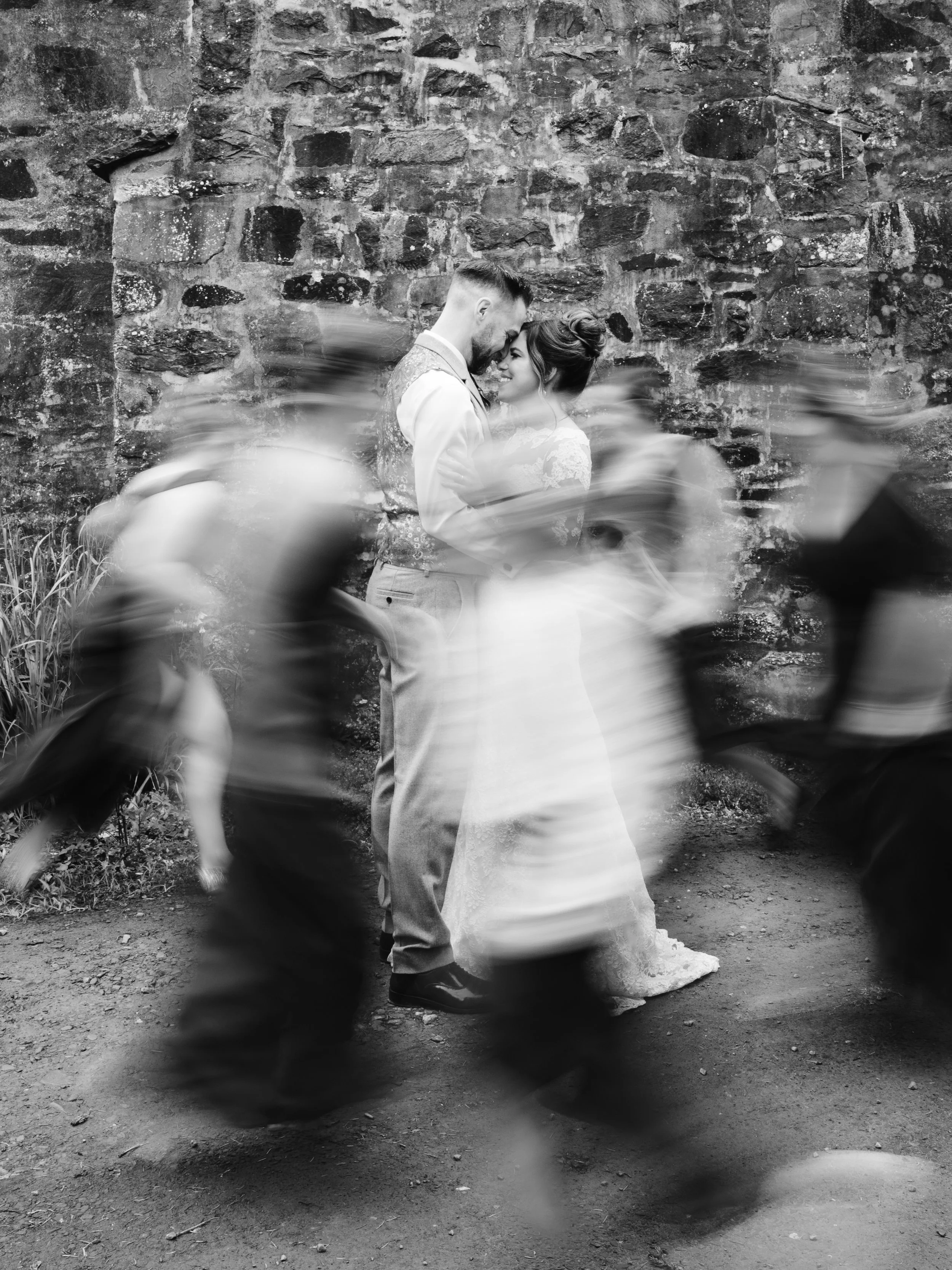 A black-and-white photo of a couple in wedding attire, embracing and touching foreheads against a rustic brick wall. Blurred figures of wedding guests are moving in front of the camera in the foreground.