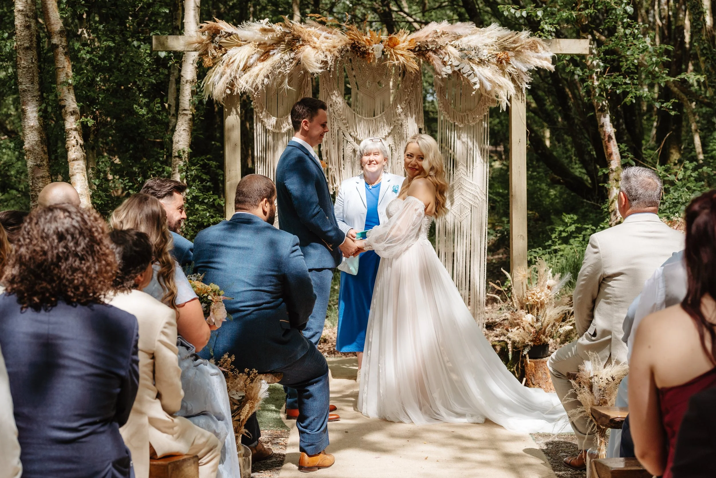 A couple gets married outdoors during daytime at a rustic wedding ceremony under a decorated wooden arch with drapes and floral arrangements. Guests seated on either side watch as the bride and groom exchange vows.