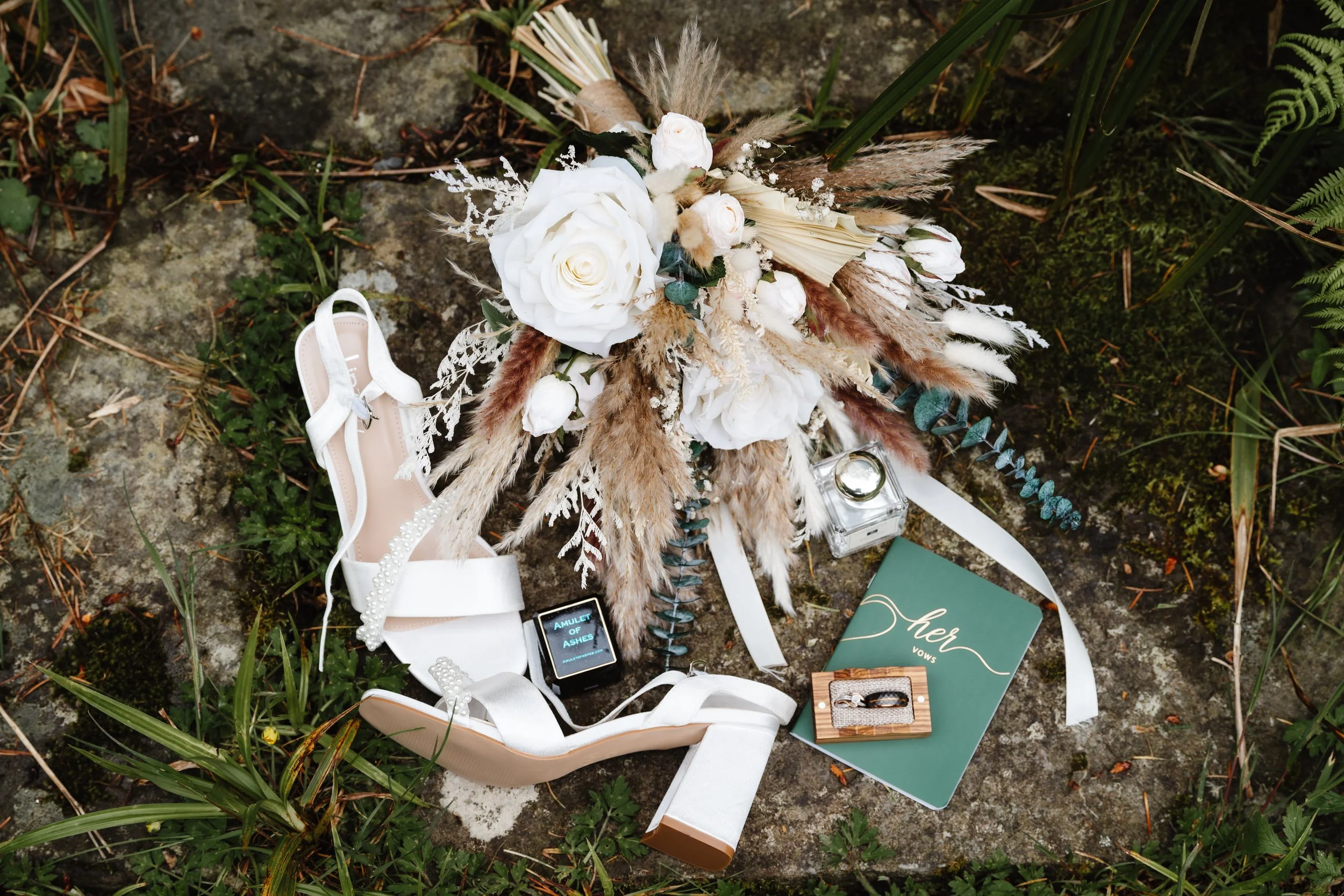 A wedding bouquet with white roses, dried pampas grass, and assorted greenery, lying on the ground surrounded by grass and moss. Nearby, a pair of white bridal shoes with pearl embellishments, a glass perfume bottle, a book titled "Her Vows," a small