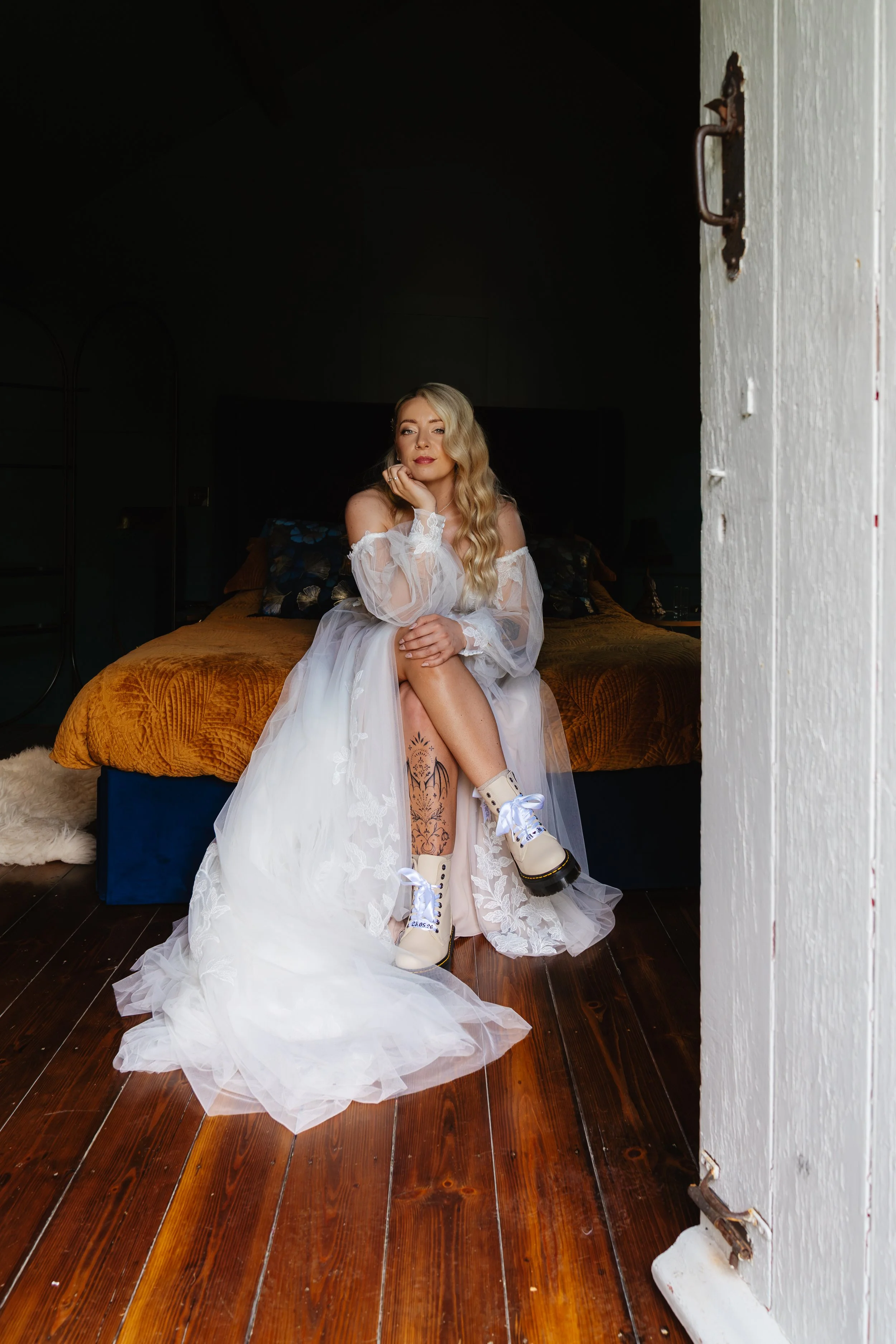 A woman in a white wedding dress sits on a bed with a yellow-orange blanket, wearing white boots with black soles and laces, with a tattoo on her left leg. She has long blonde hair styled in loose waves. The room has dark walls and wooden floors, wit