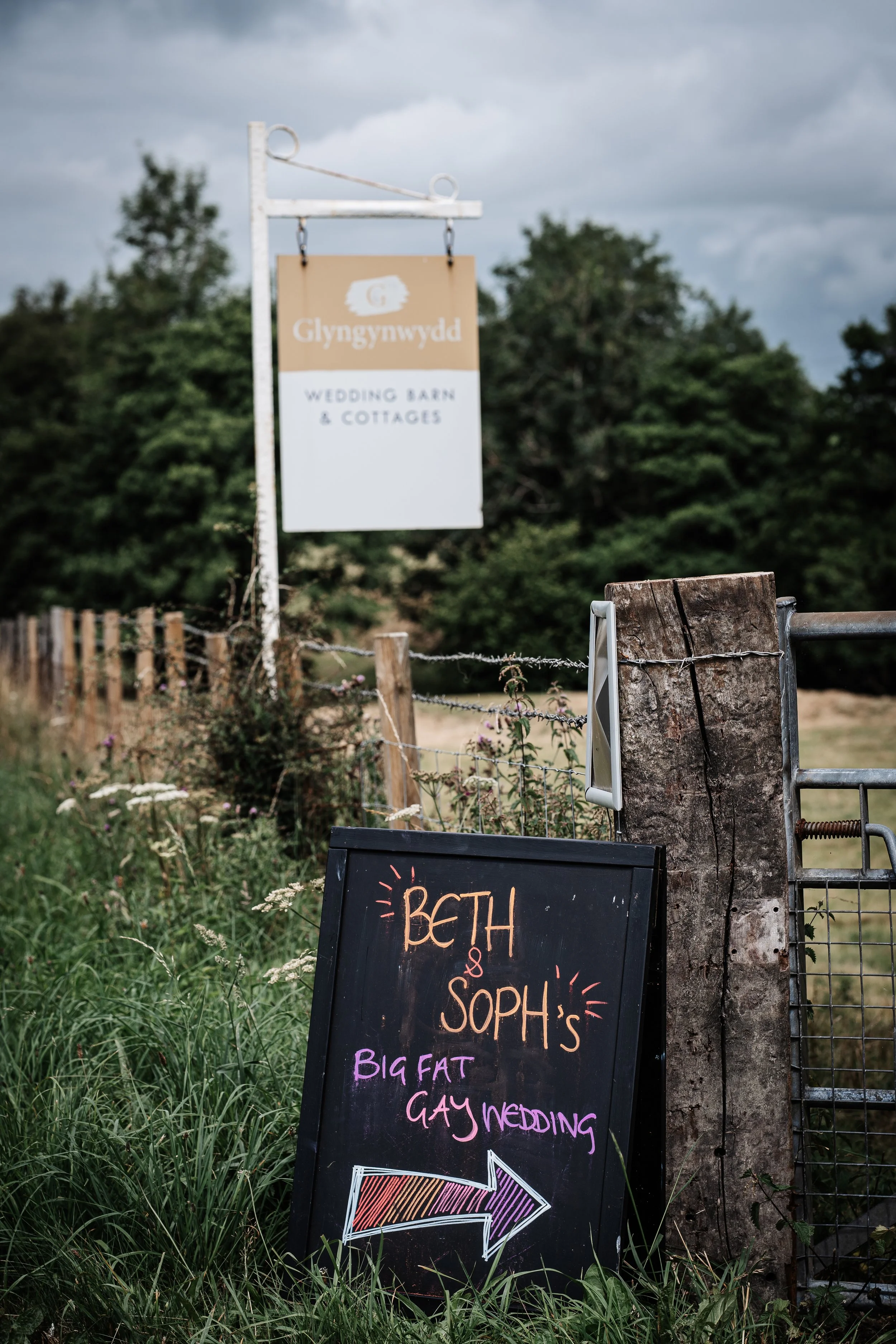 A blackboard sign with colorful chalk writing advertising Beth and Soph's big fat gay wedding, with an arrow pointing to the right. Behind it is a gate and wooden post, and further back is a sign with the Welsh name 'Glyngynwydd' indicating a wedding