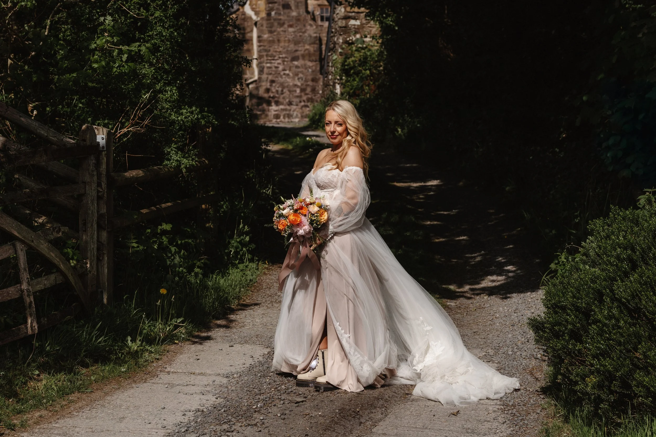 A bride wearing a white wedding gown and holding a bouquet of flowers stands on a dirt pathway in a lush green garden with a stone tower in the background.