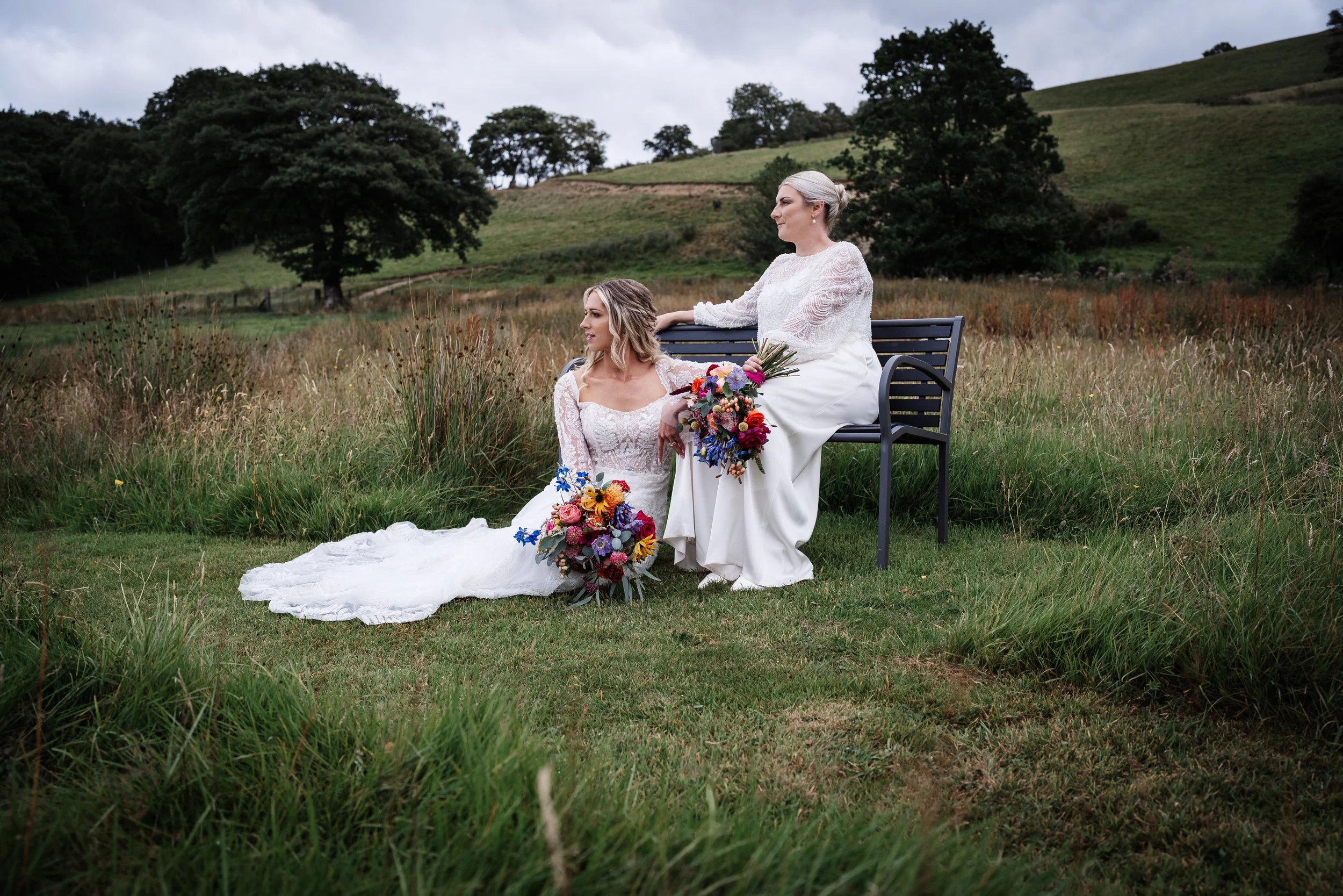 Two women in white wedding dresses, one sitting on a black bench and the other sitting on the grass, holding bouquets of colorful flowers, in an outdoor green field with trees and hills in the background.