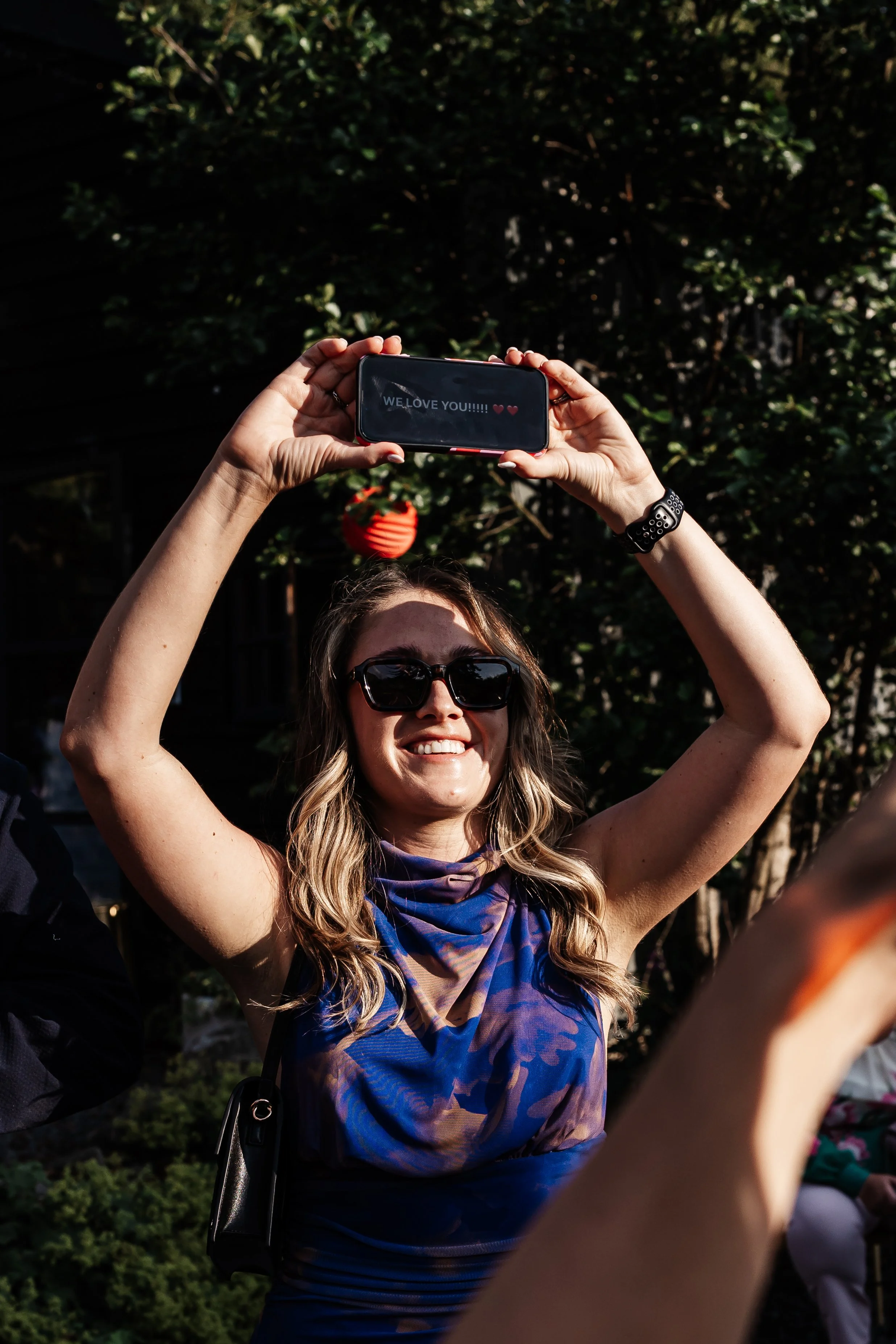 Woman with long hair, sunglasses, and a blue patterned scarf, smiling as she takes a selfie with her phone, displaying a message that says "WE LOVE YOU!!!!! ❤️❤️".