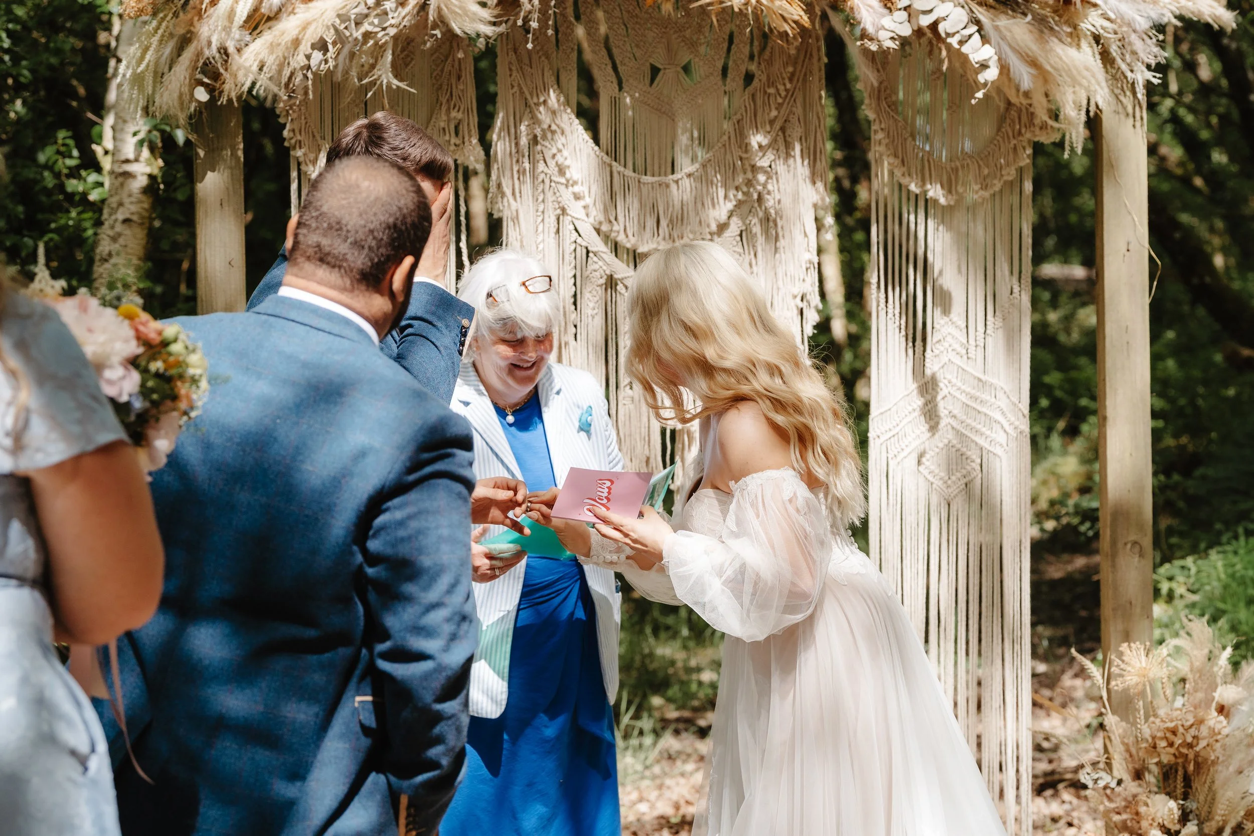 A wedding ceremony outdoors with a young girl dressed as a bride, holding a certificate or certificate of marriage, standing under a rustic woven canopy. Adults, including an elderly woman with glasses and gray hair, and a man in a blue suit, are pre