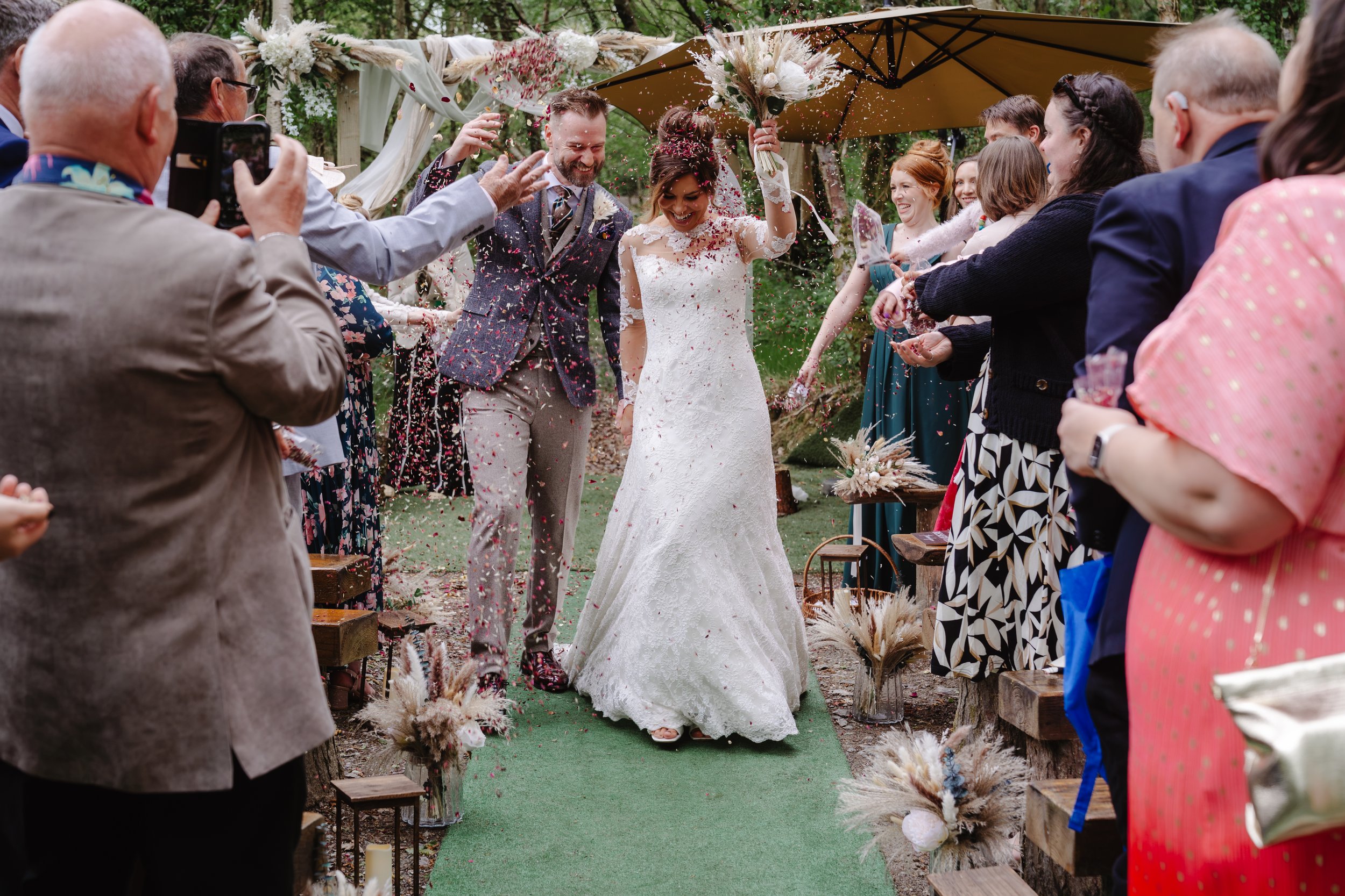 A bride and groom are walking down an outdoor aisle at their wedding, surrounded by family and friends, with confetti being thrown and a decorated archway above.