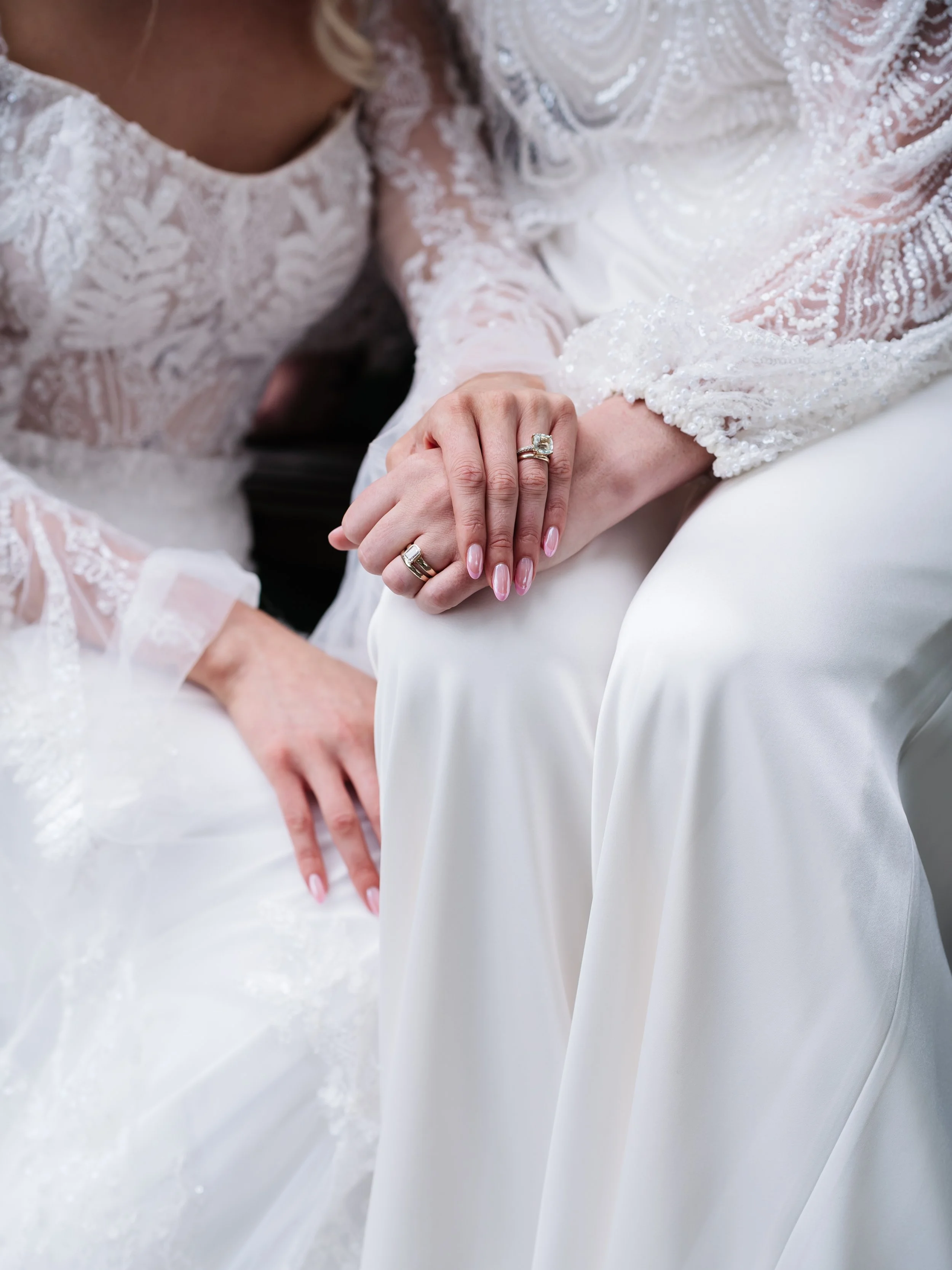 Two women in wedding dresses holding hands, showing rings on their fingers.