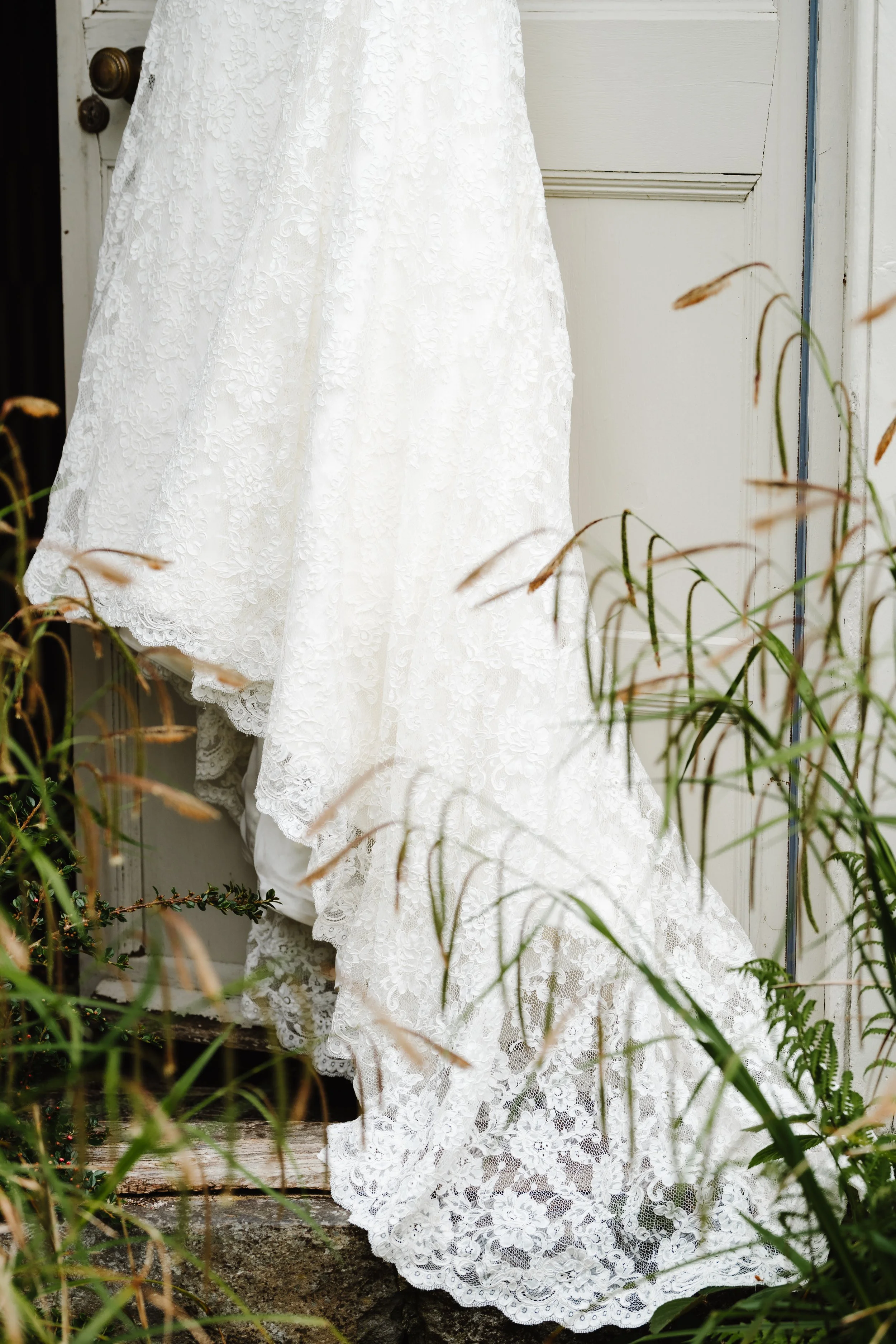White lace wedding dress hanging outside near a white door, with green plants and grass around
