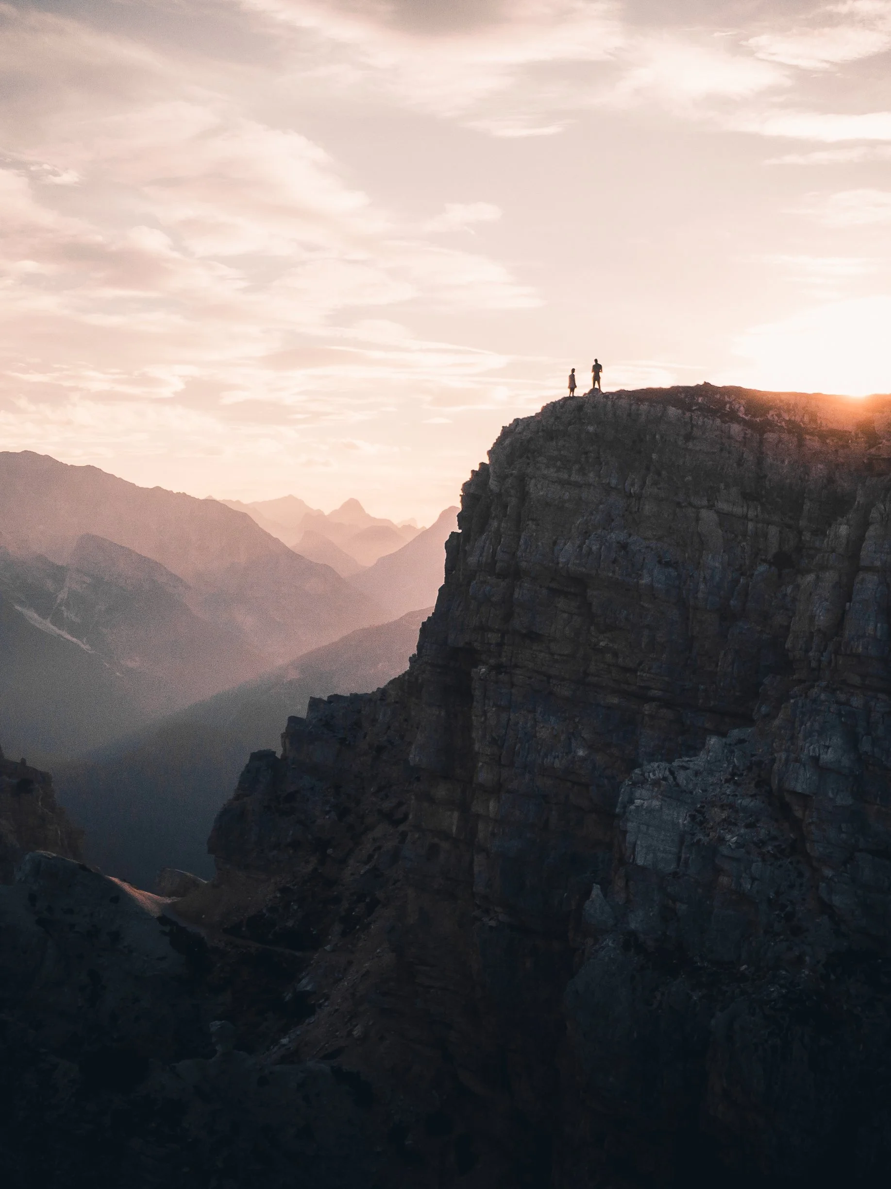 Two people standing on a rocky mountain peak during sunset, overlooking distant mountain ranges and a cloudy sky.