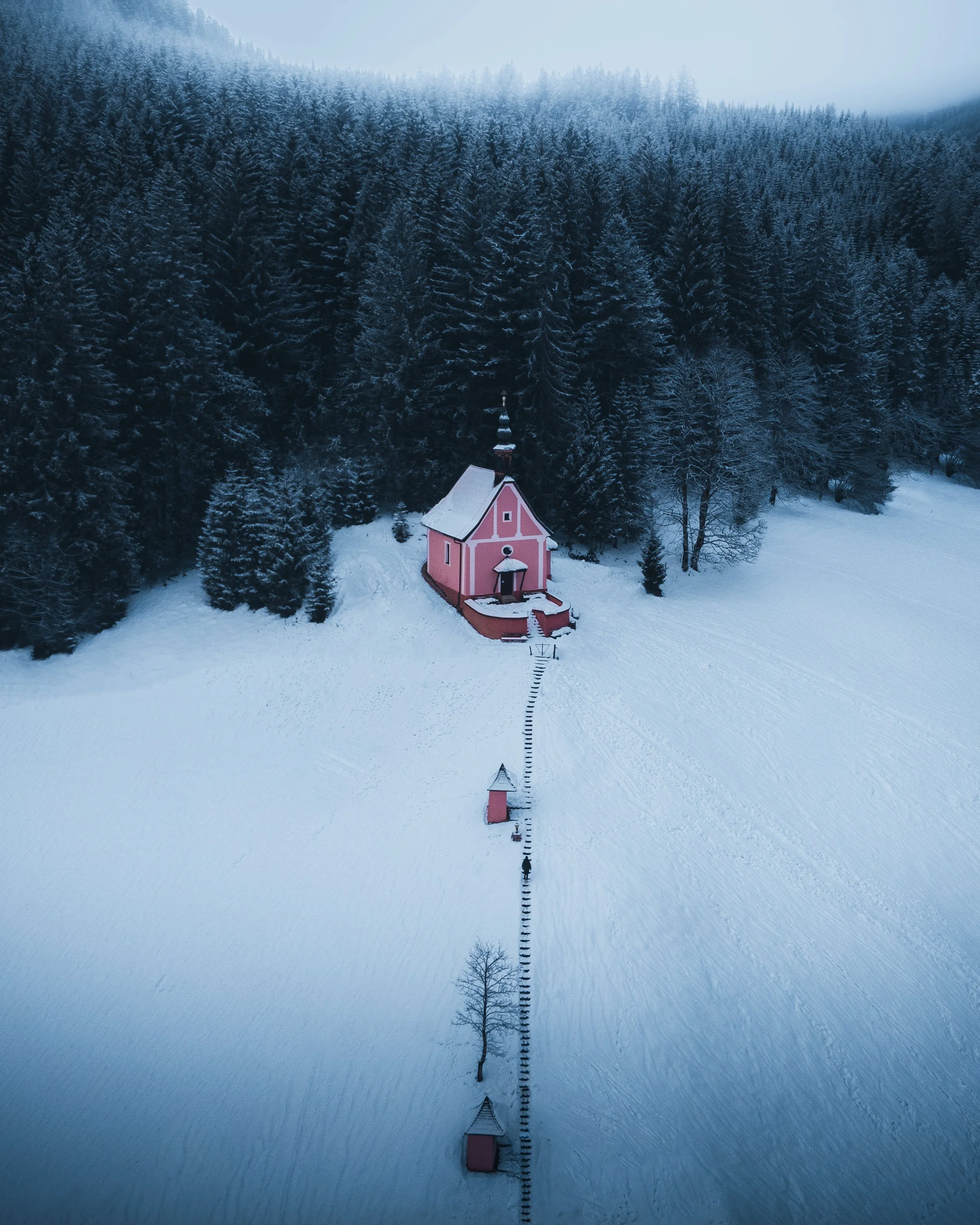 A snow-covered landscape featuring a small pink church with a tall steeple and a person walking on a trail leading to the church. The background is a dense forest of snow-dusted evergreen trees, with mountains shrouded in fog in the distance.