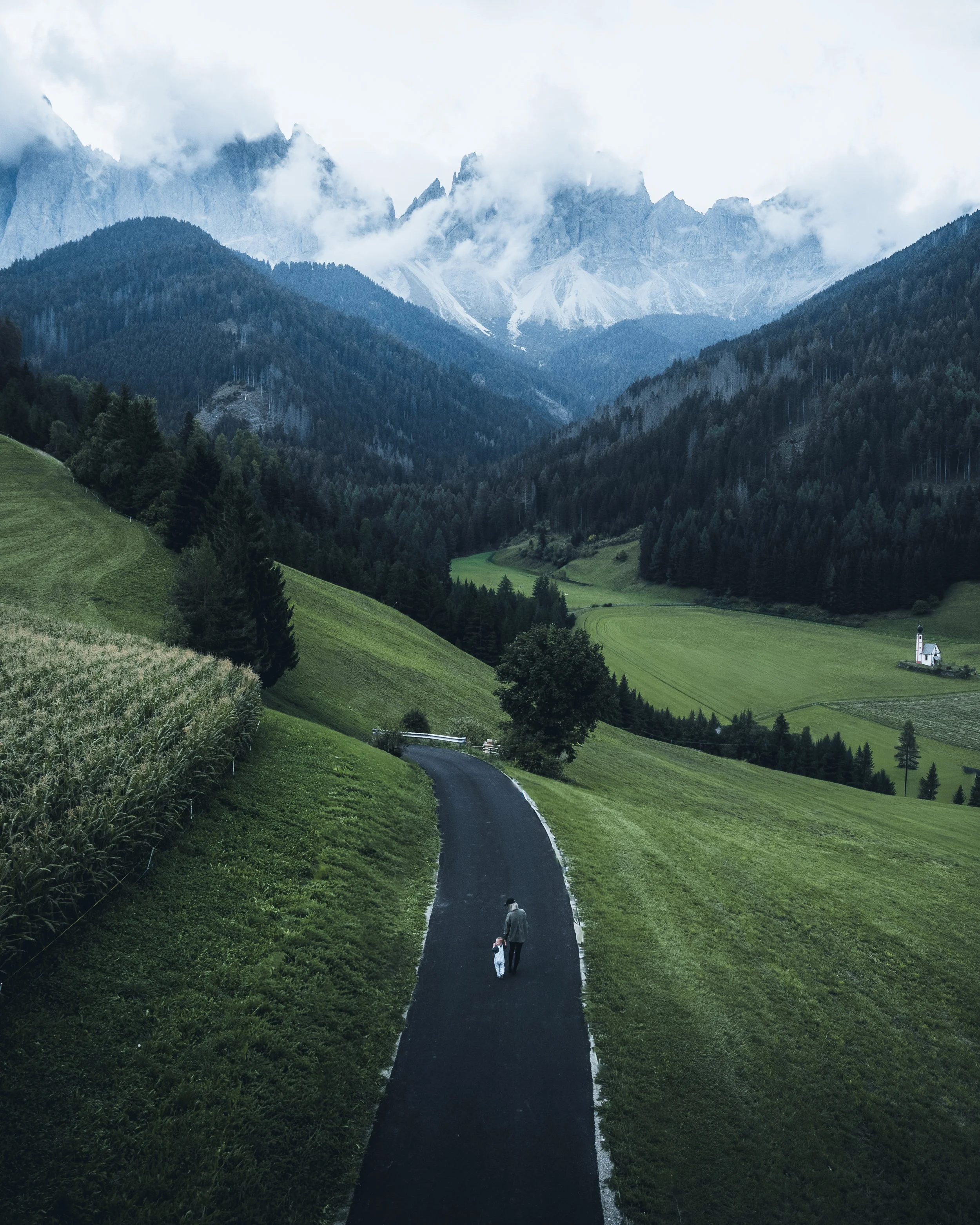 A winding paved road through lush green hills with a person and child walking, mountains with clouds in the background under an overcast sky.