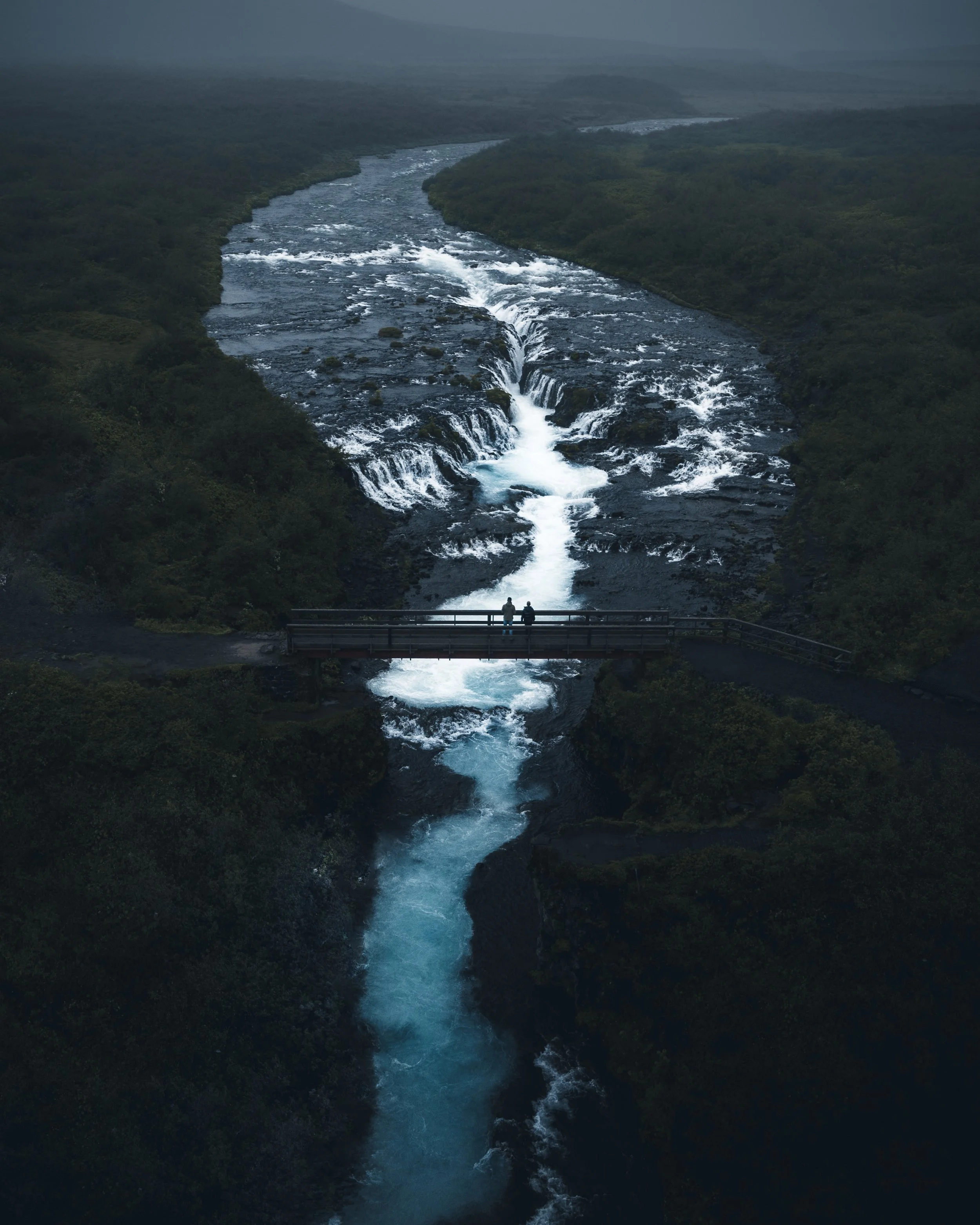 Two people standing on a bridge overlooking a wide river with waterfalls, lush green banks, and rolling hills in the background under overcast sky.