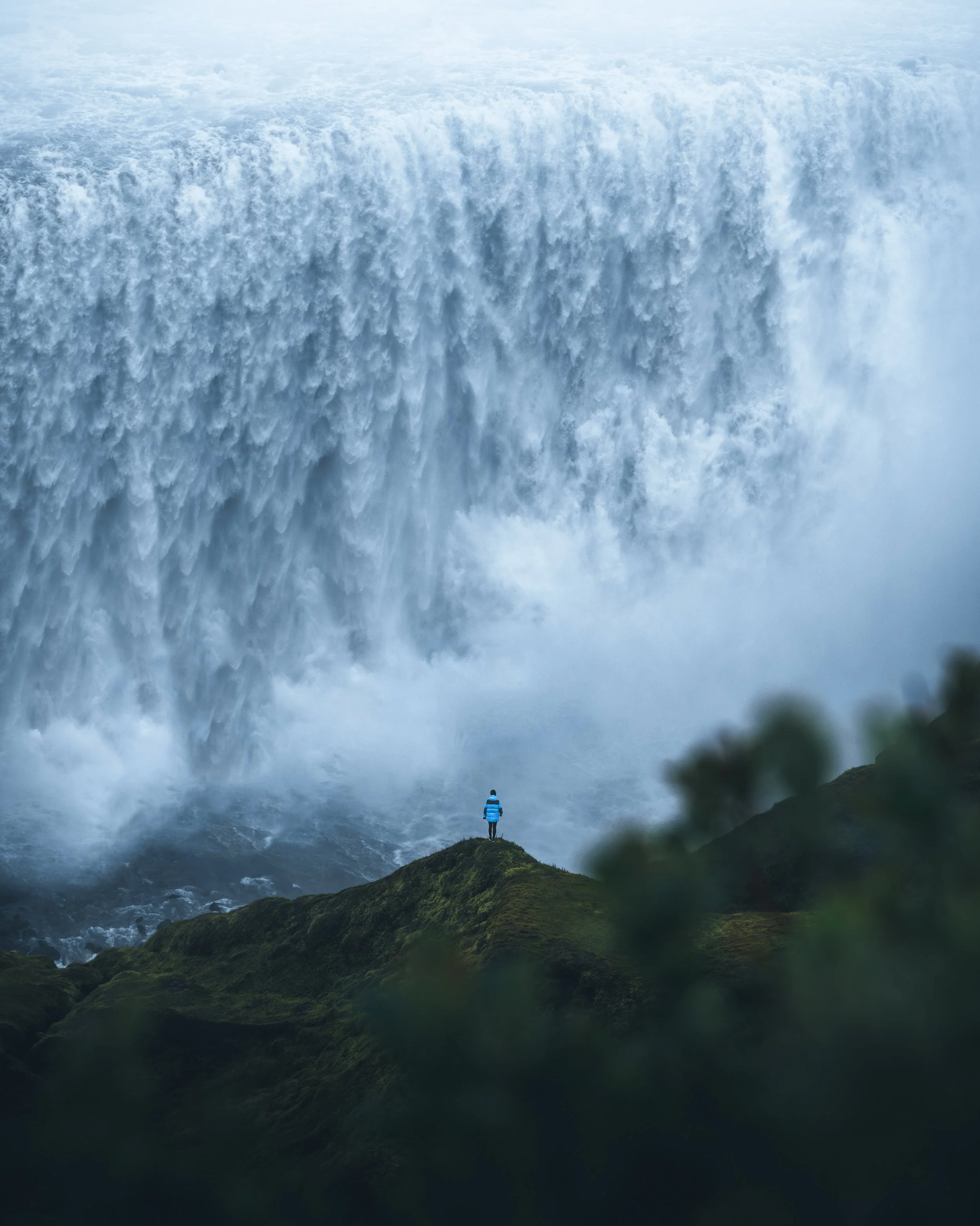 A person dressed in a blue jacket standing on a patch of green grass overlooking a large, powerful waterfall with mist and spray rising from the base, surrounded by lush green hills.