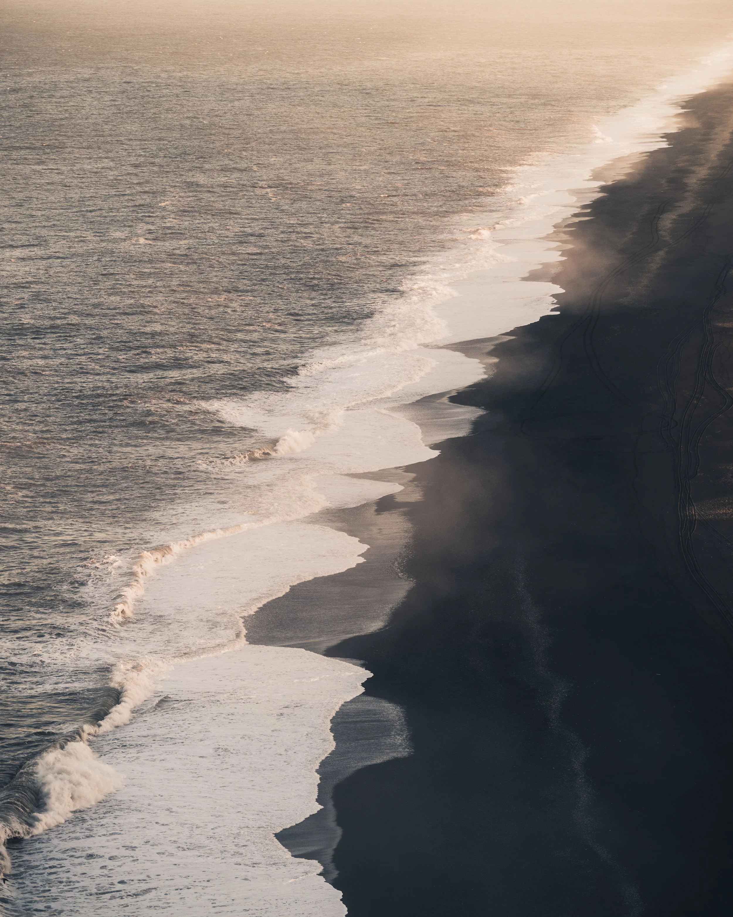 Aerial view of the ocean meeting a dark sandy beach at sunset, with waves crashing onto the shoreline.