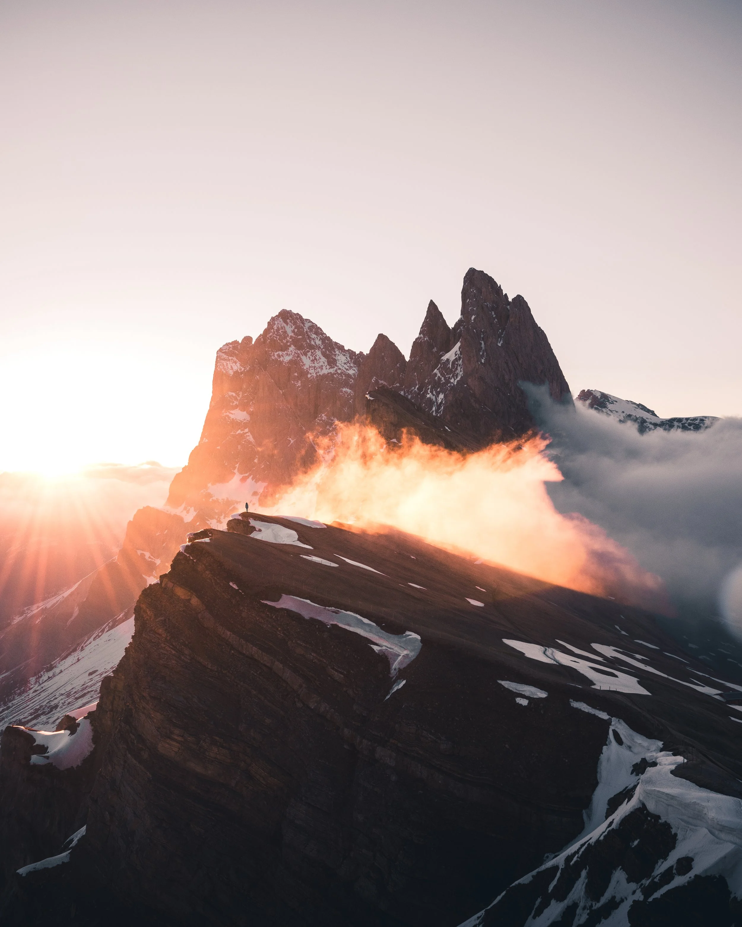Snow-capped mountain peaks at sunrise with a person standing on a ridge, surrounded by clouds and mist.