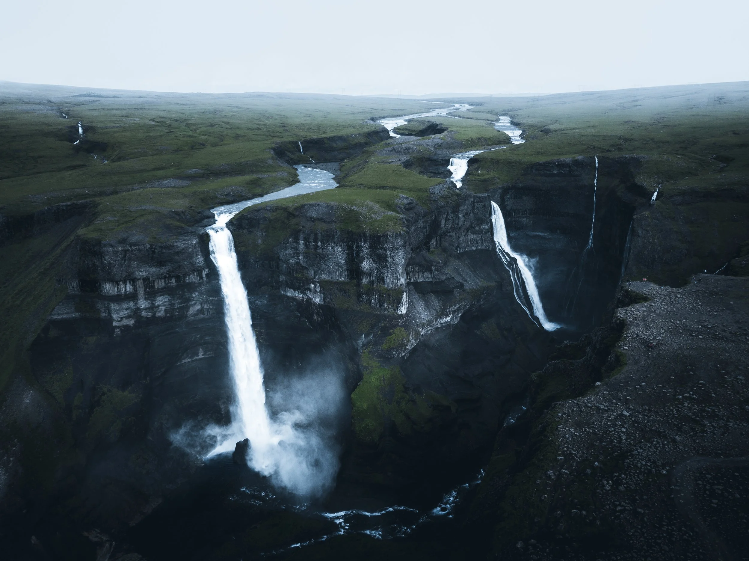 Multiple waterfalls cascading down rocky cliffs into a river below in a lush green landscape under an overcast sky.