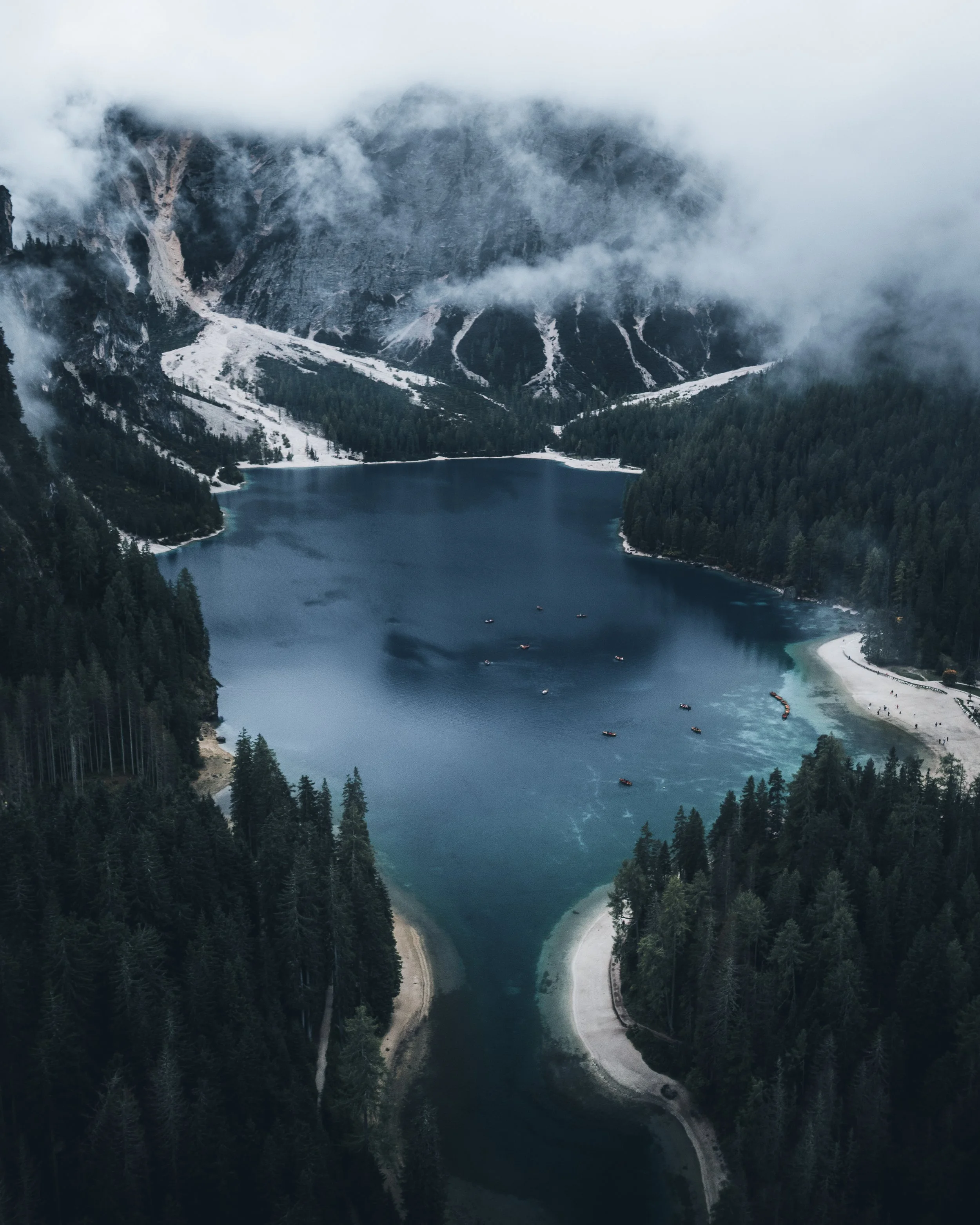 A mountain lake surrounded by dense evergreen trees and a winding dirt road, with mist-covered mountains in the background.