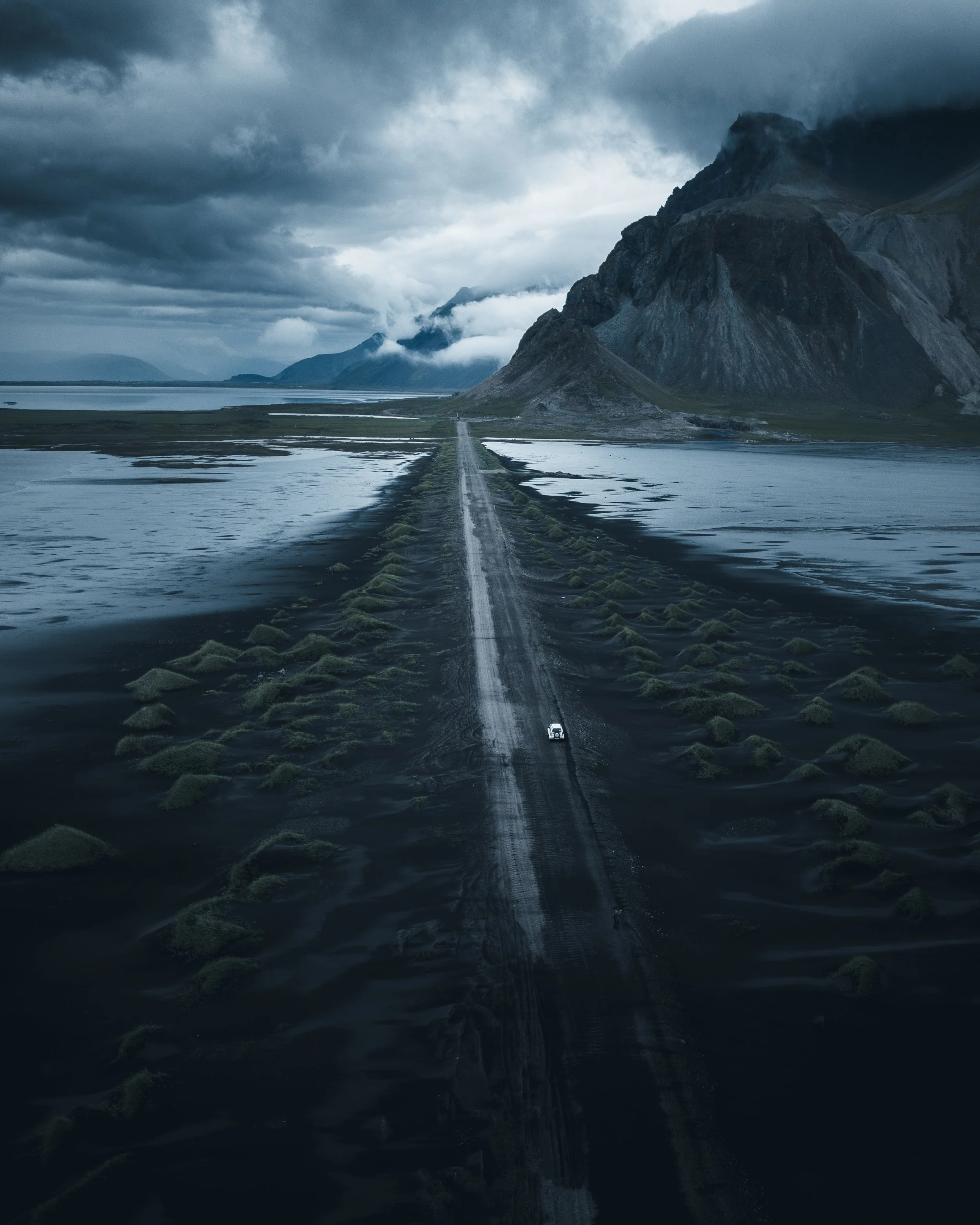 A narrow road stretches into the distance, flanked by water on both sides, leading towards a mountain range under a dark, cloudy sky.