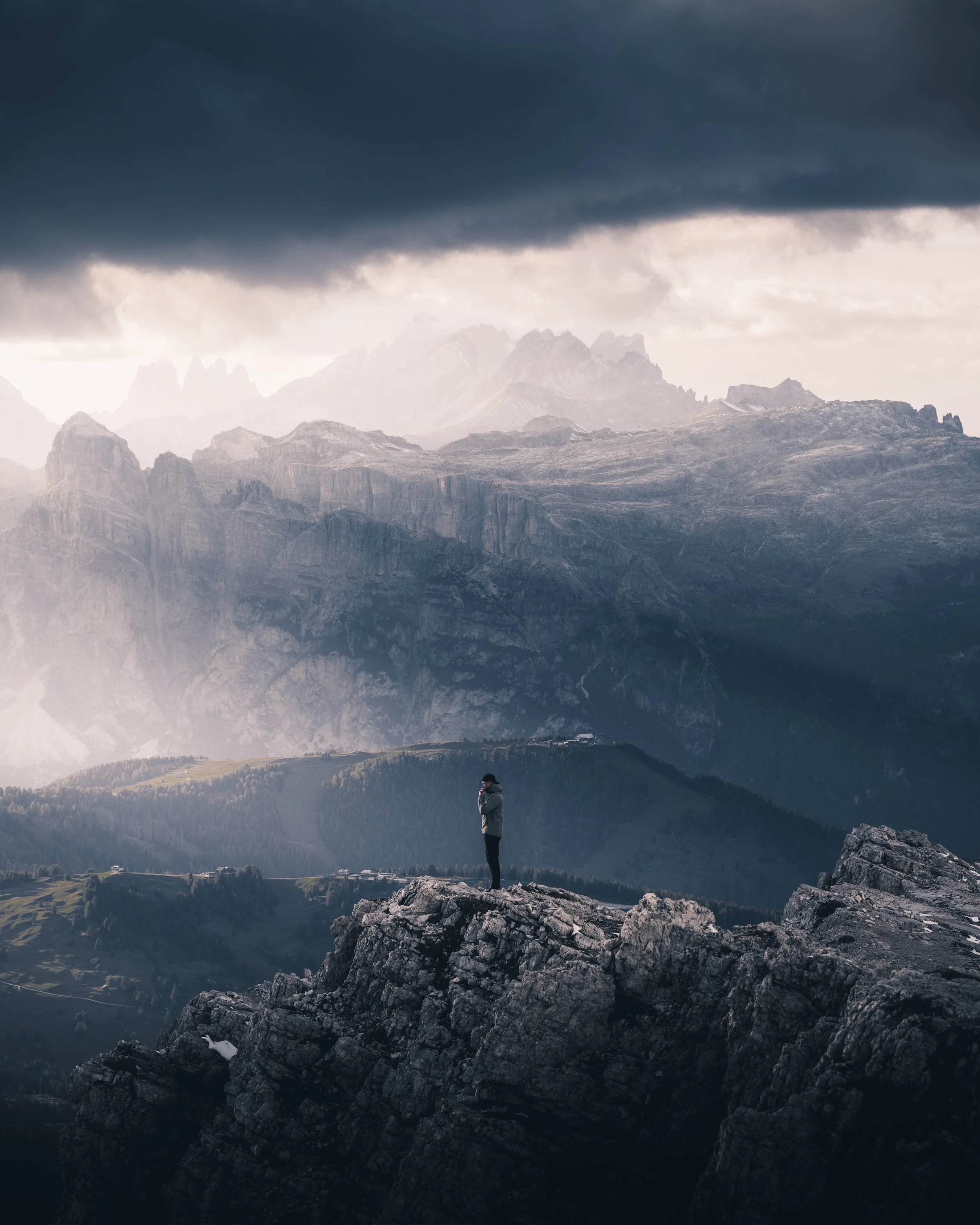 A person standing on a rocky outcrop during daytime, looking at a mountain range with dark, stormy clouds overhead.