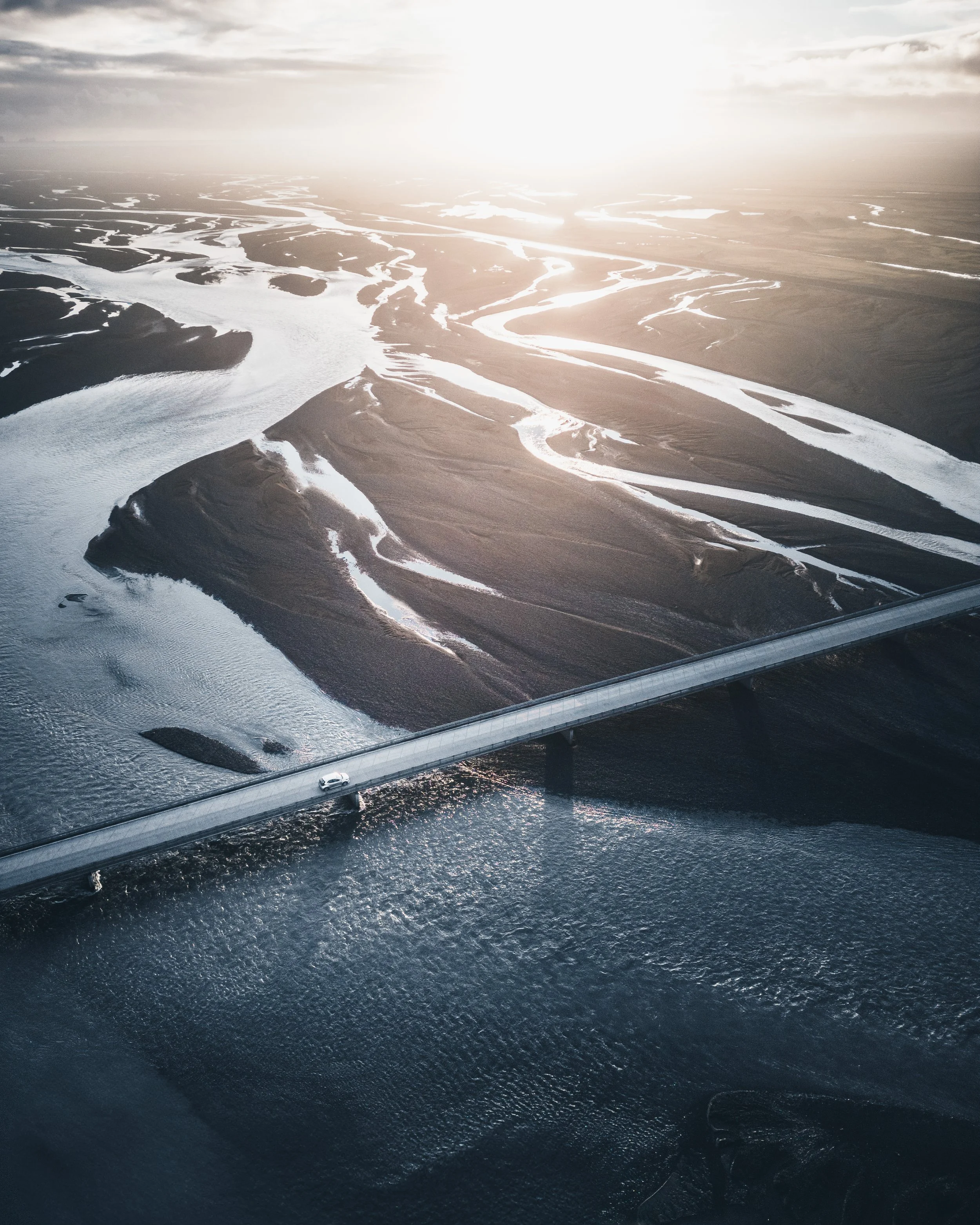 Aerial view of river channels and wetlands with a bridge crossing over water, under a cloudy sky at sunset.