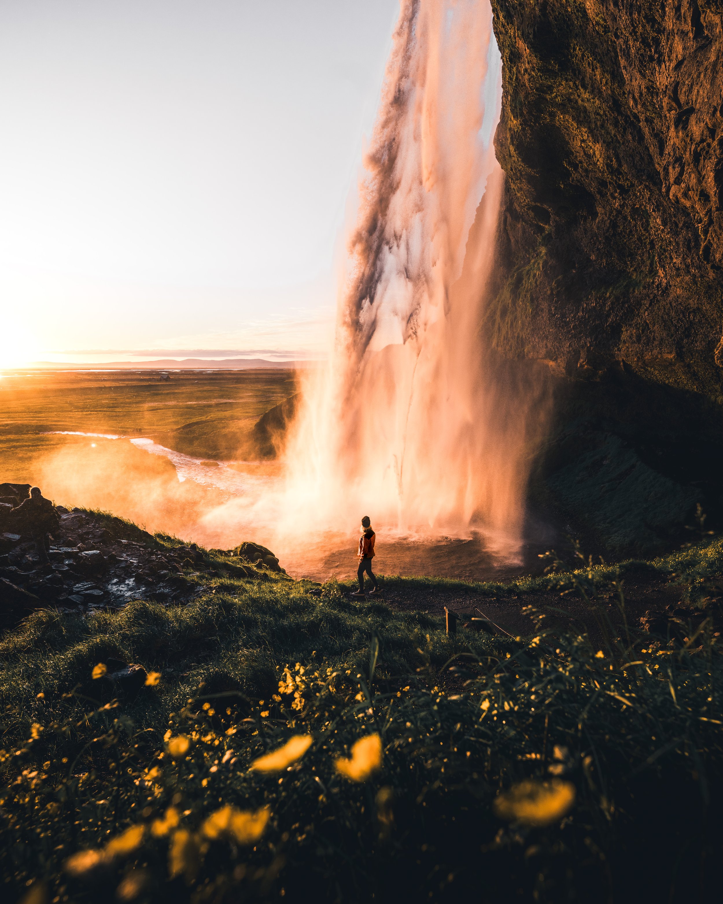 A person standing near a powerful waterfall during sunset or sunrise, surrounded by lush greenery and yellow flowers in the foreground.